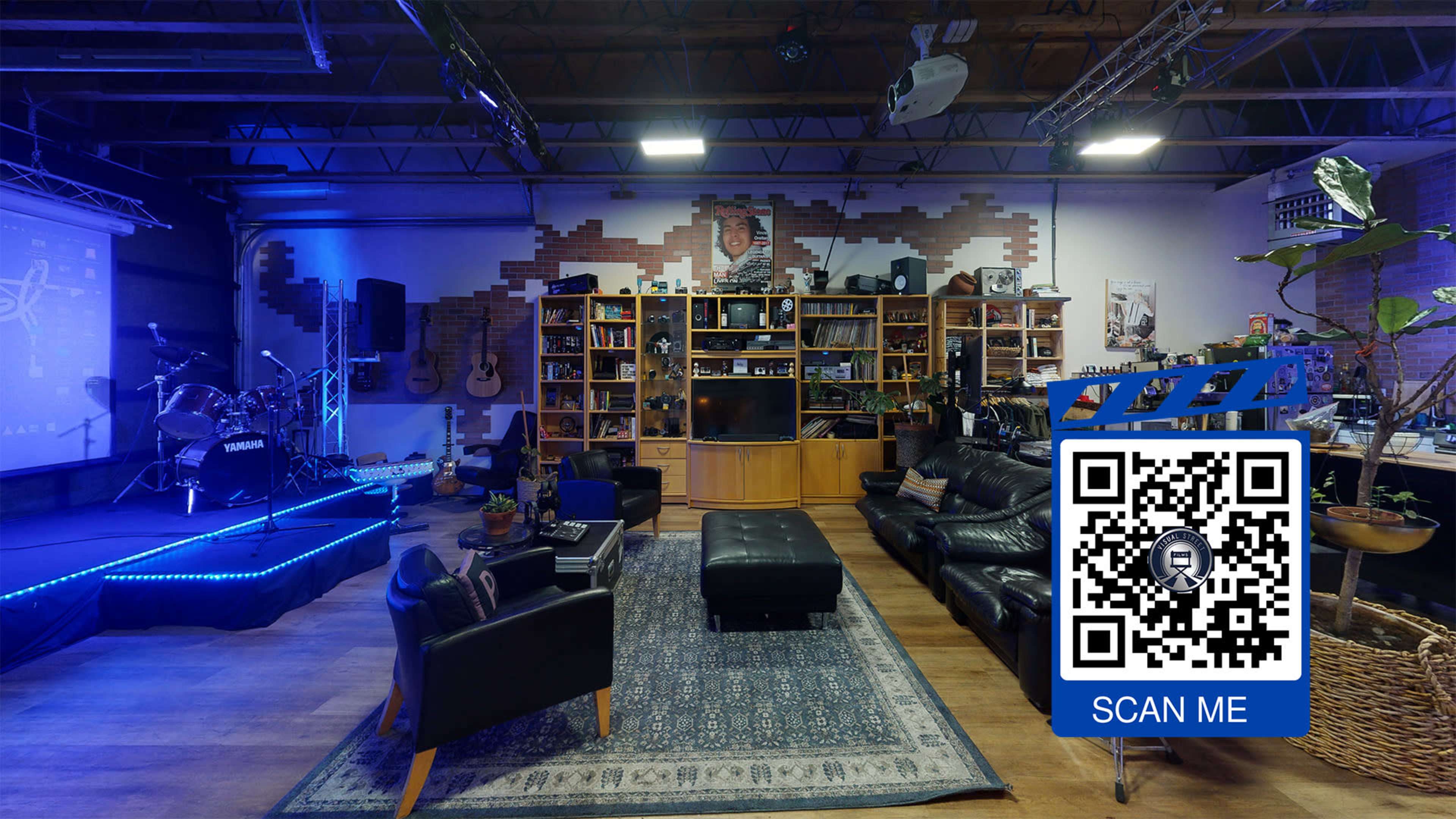 A cozy music studio or rehearsal space with a stage featuring musical instruments, a large bookshelf filled with items, and seating areas with black leather furniture.