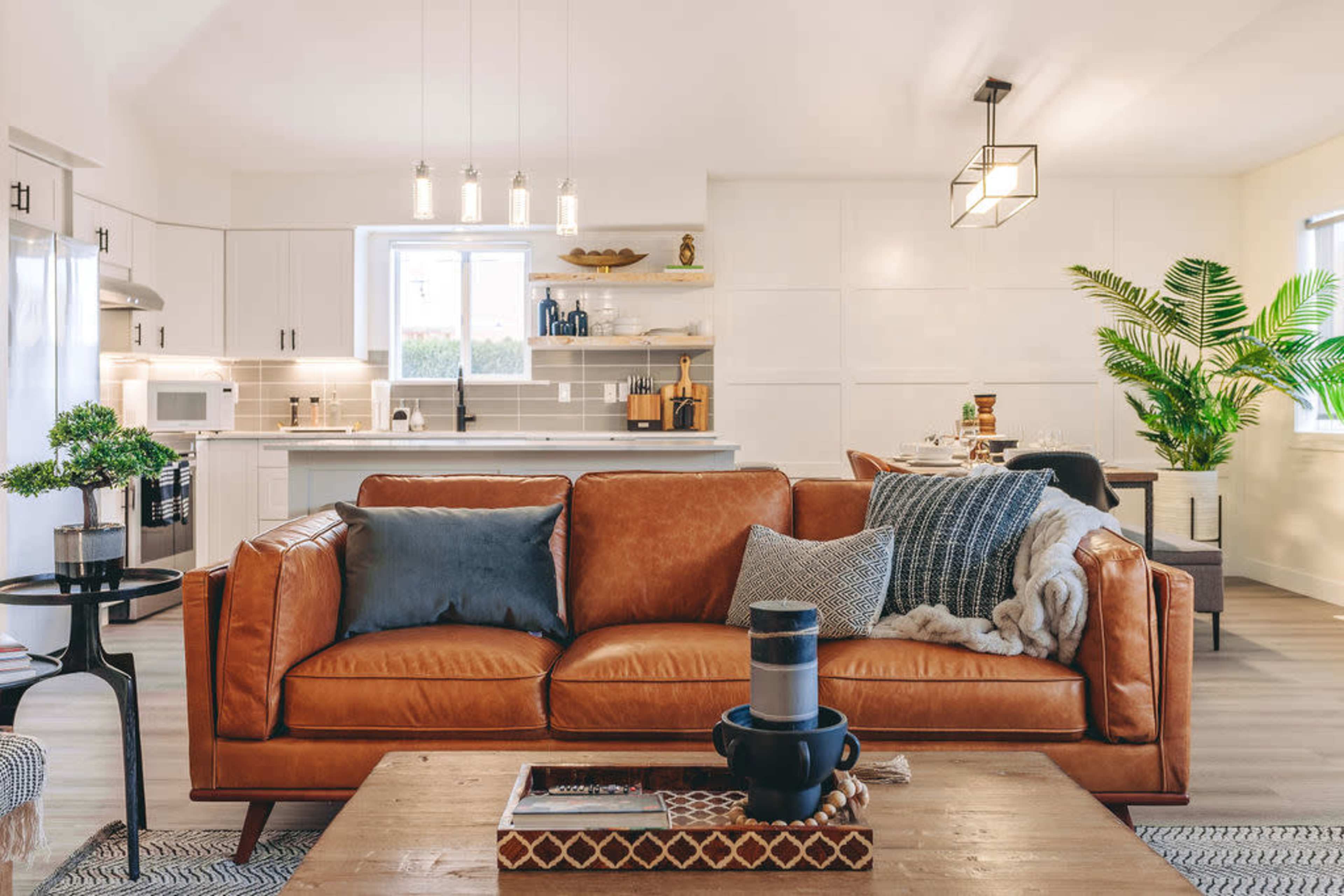 A modern living room features a brown leather sofa with decorative pillows, a wooden coffee table, and an open kitchen space in the background.