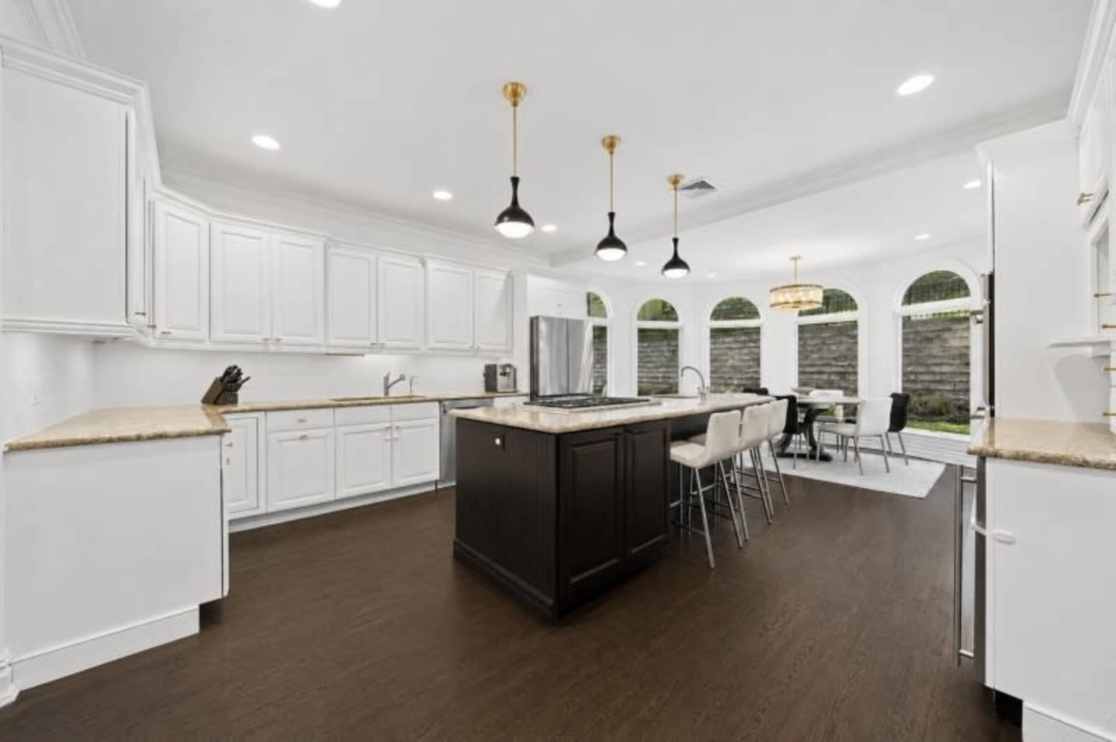 The kitchen features white cabinetry, a large central island with a dark wood finish, and a dining area with large windows that overlook a stone wall garden.