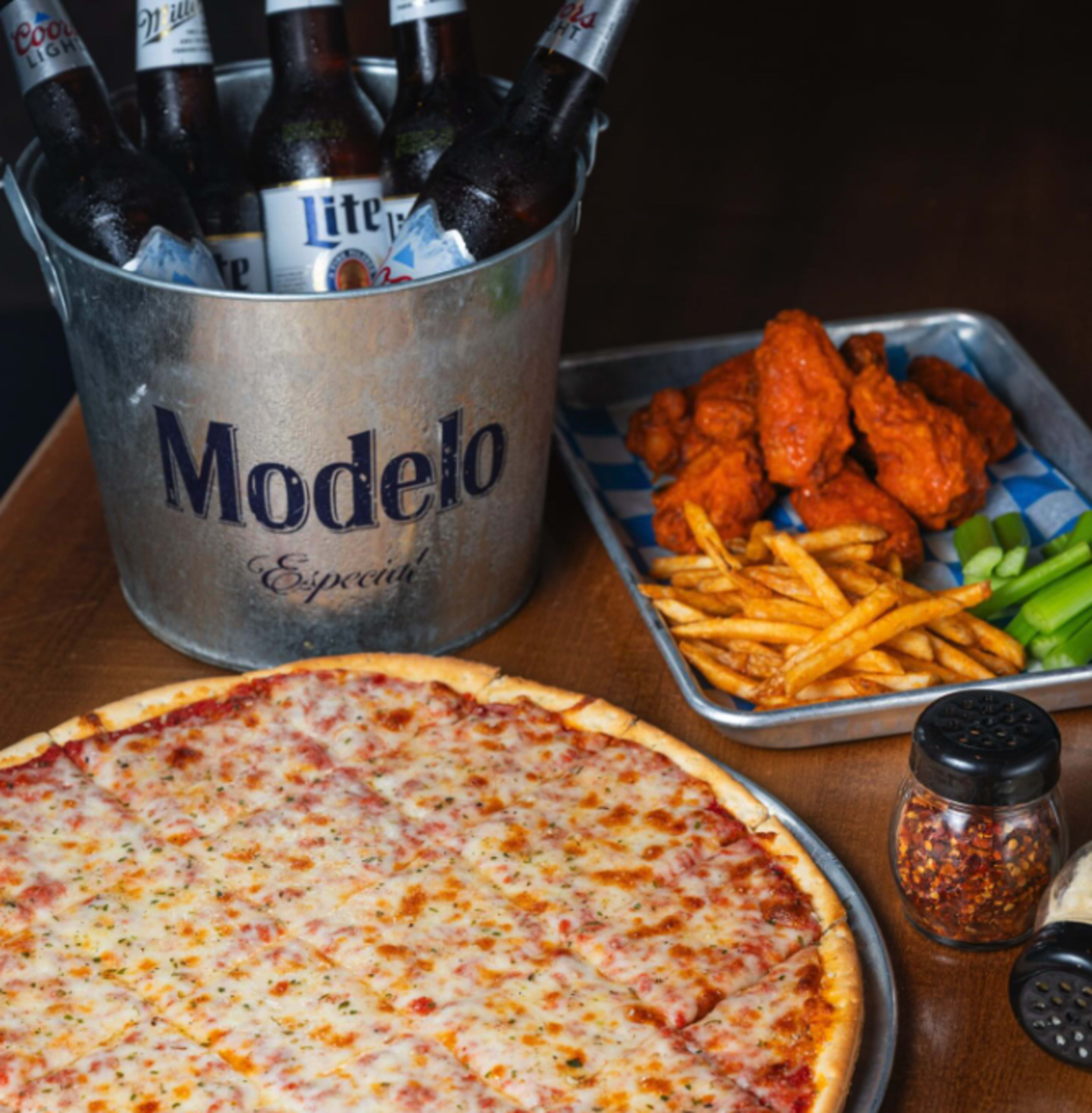 The image shows a table setup with a bucket of beer bottles, a tray of chicken wings with fries and celery, and a large cheese pizza.