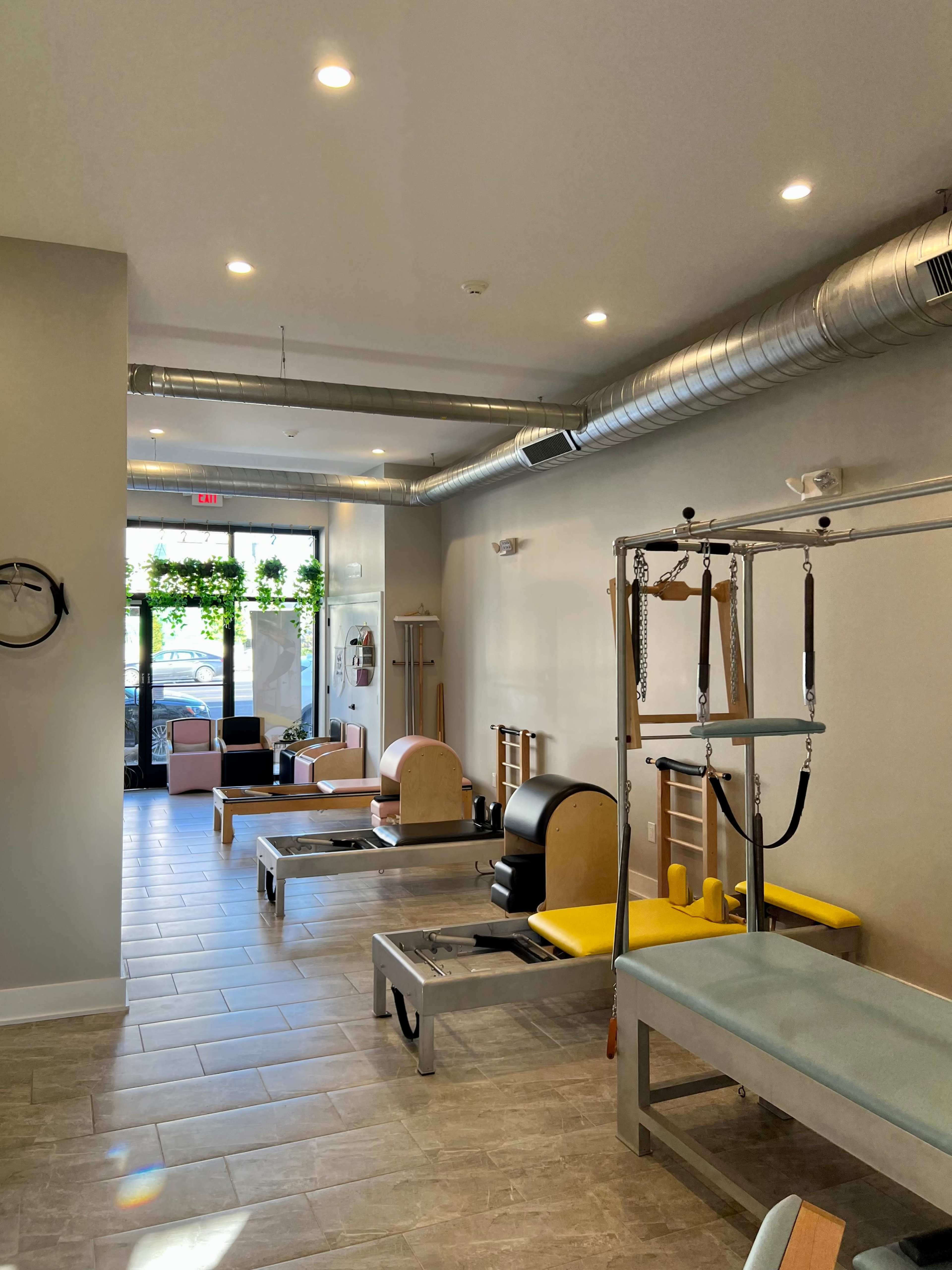 A Pilates studio featuring reformer machines, wall-mounted equipment, and large windows with natural light.