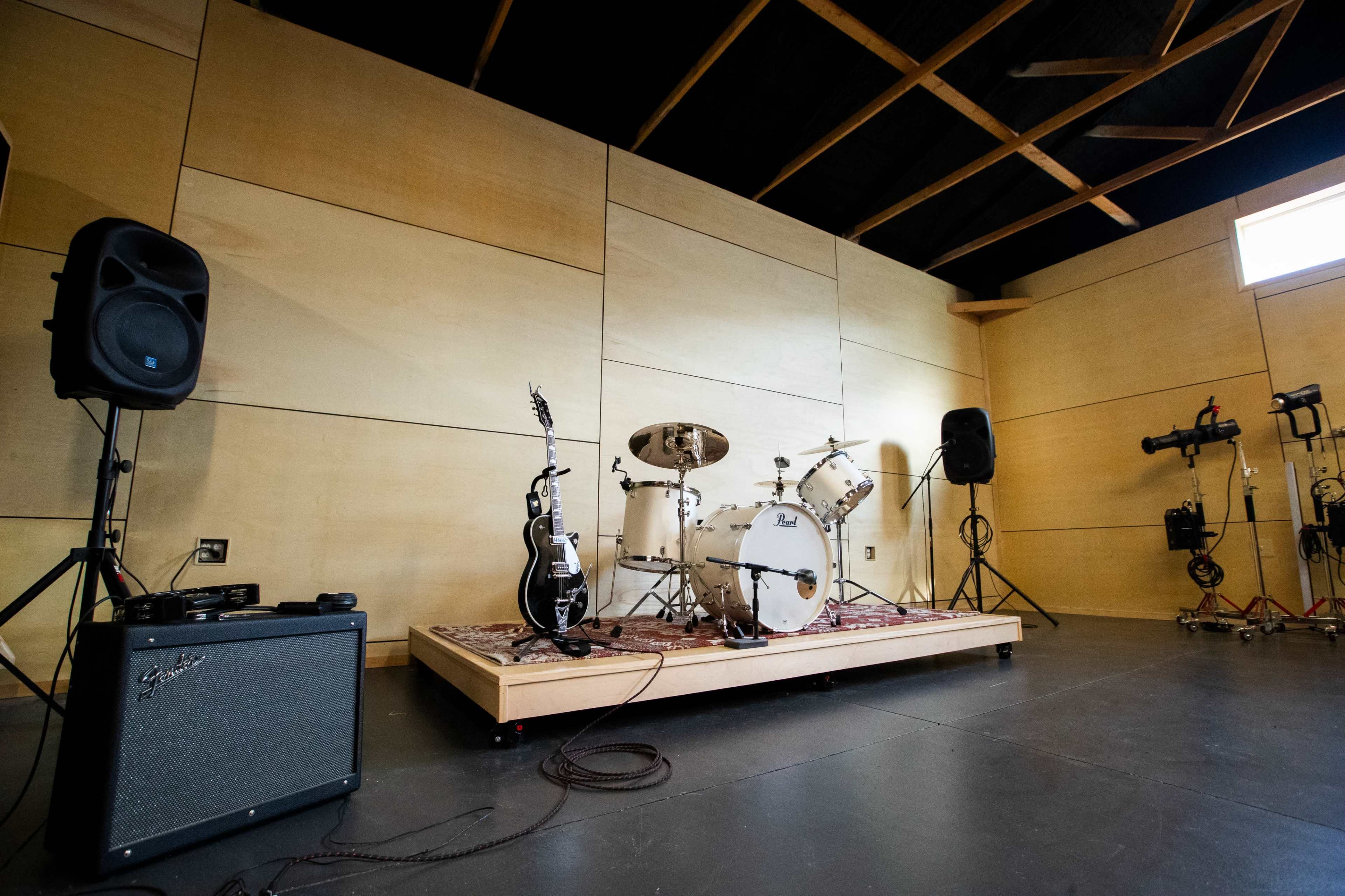 The image shows a music rehearsal space featuring a drum set, an electric guitar, amplifiers, and speakers on a raised platform with wooden walls.