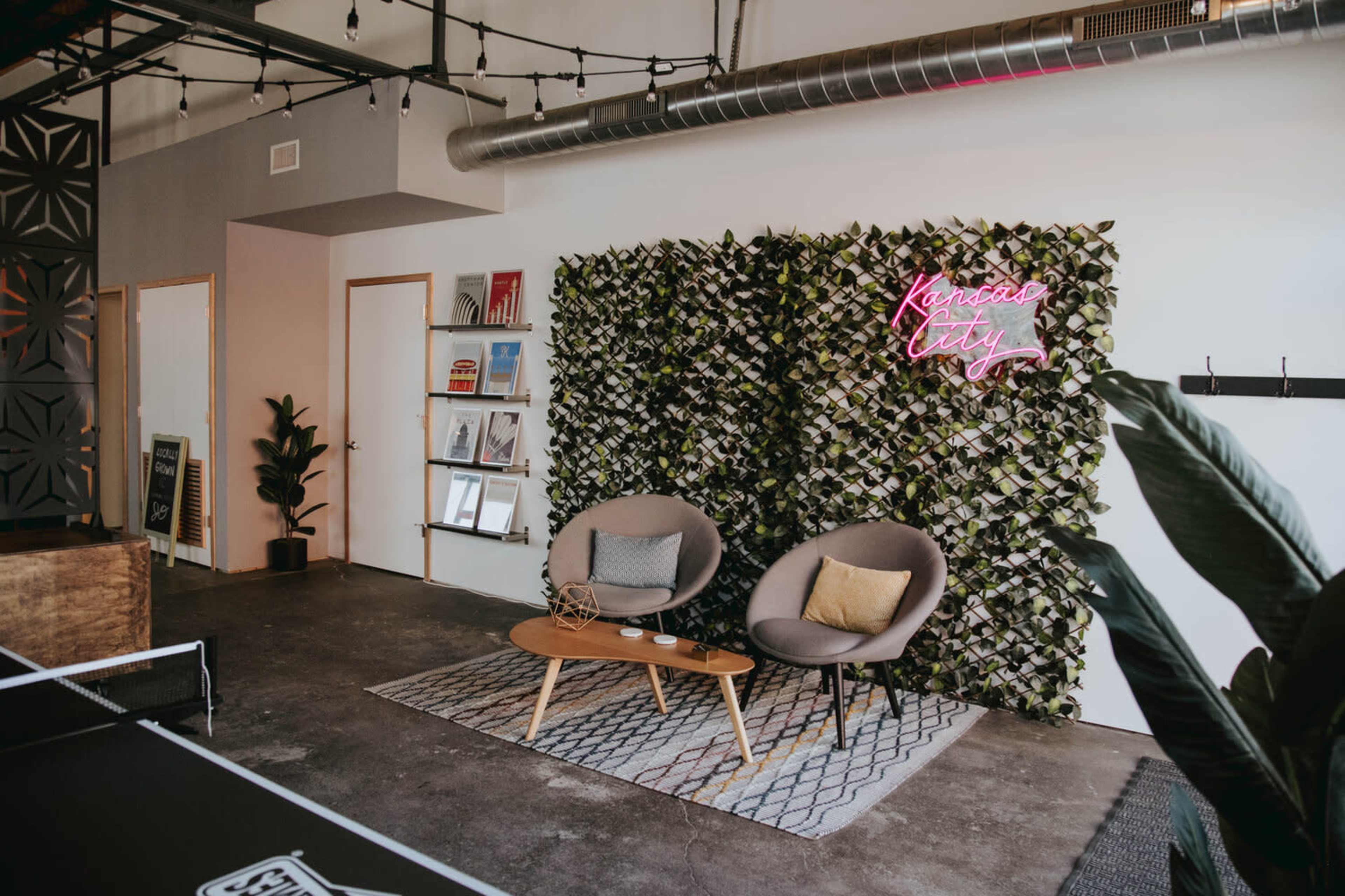 A modern indoor seating area featuring two gray chairs, a small wooden table, a plant, and a green wall with a neon sign that reads "Kansas City."
