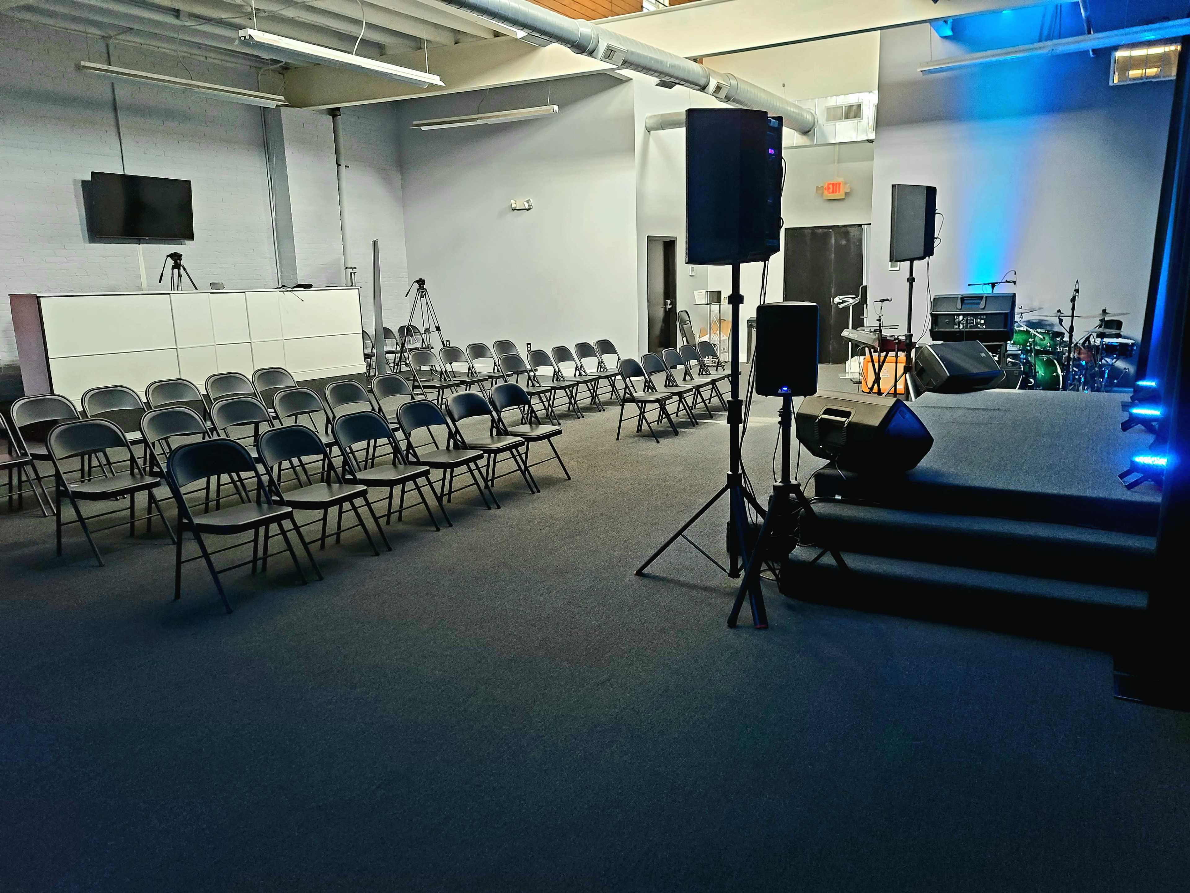 An empty auditorium set up with rows of metal chairs facing a stage, with audio equipment and a small band setup in the background.