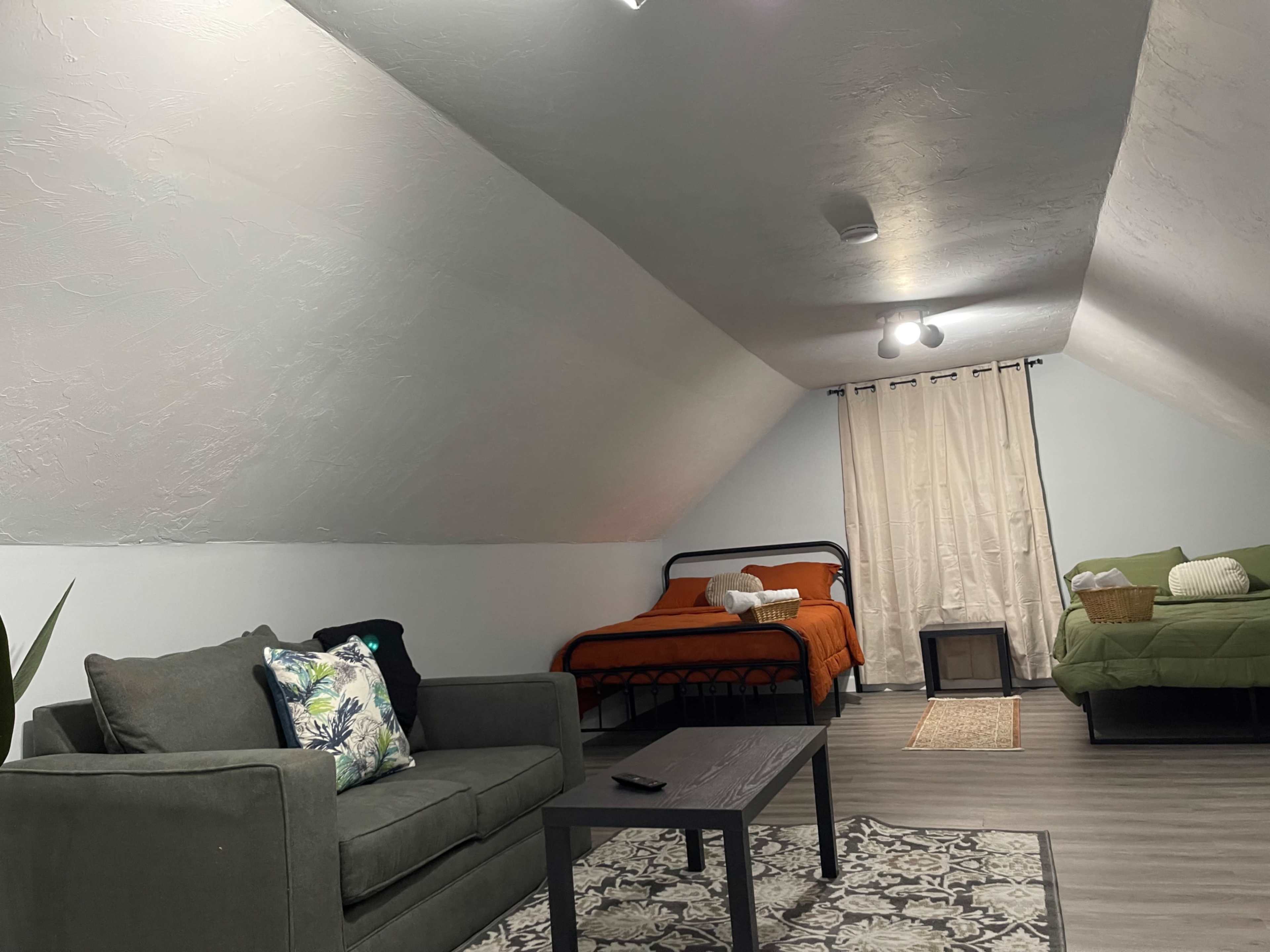 The image shows a cozy attic room with a bed, a couch, a small coffee table, and a window covered by a curtain.