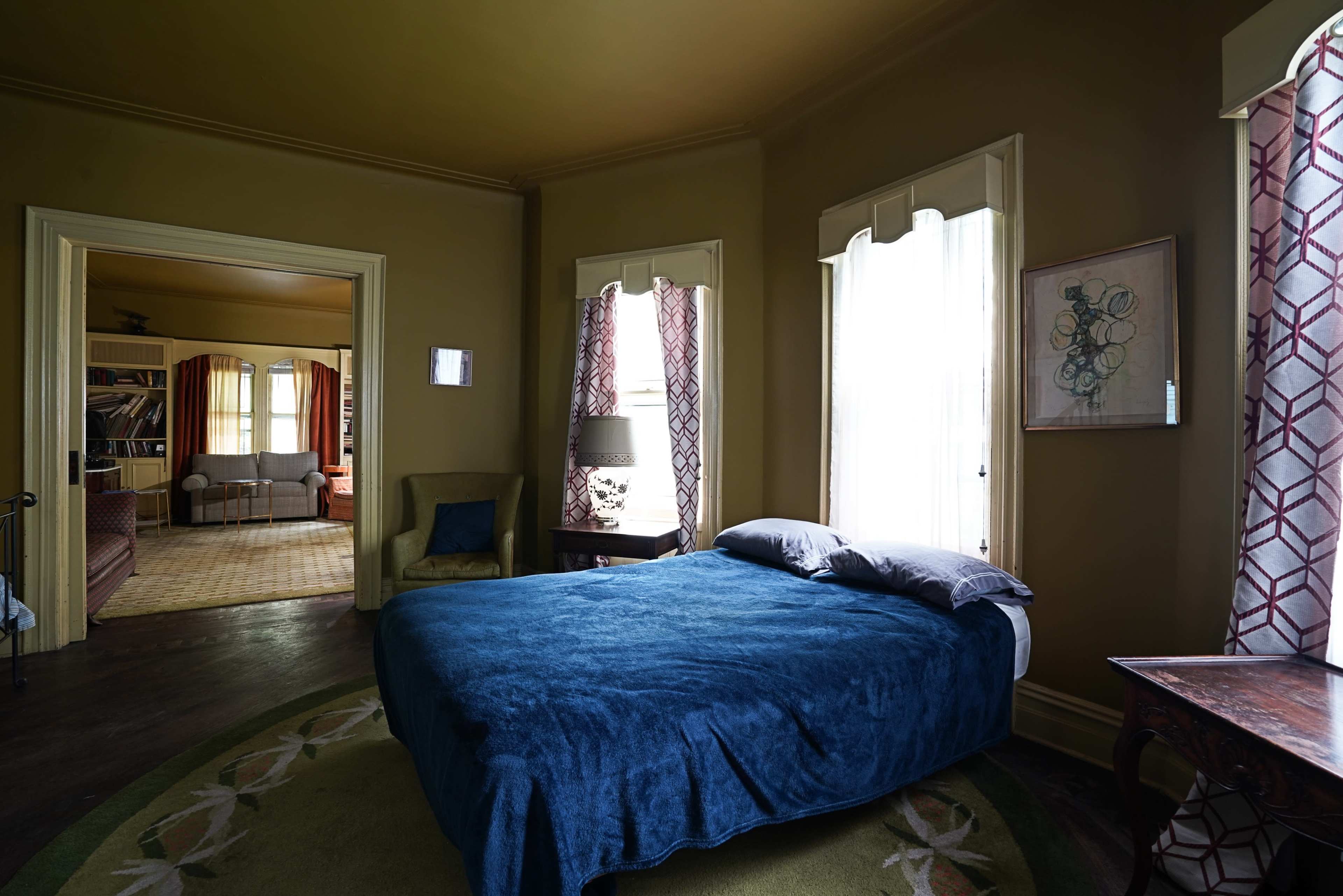 A bedroom features a bed with a blue blanket, accompanied by two windows and a view into a living area with a sofa.