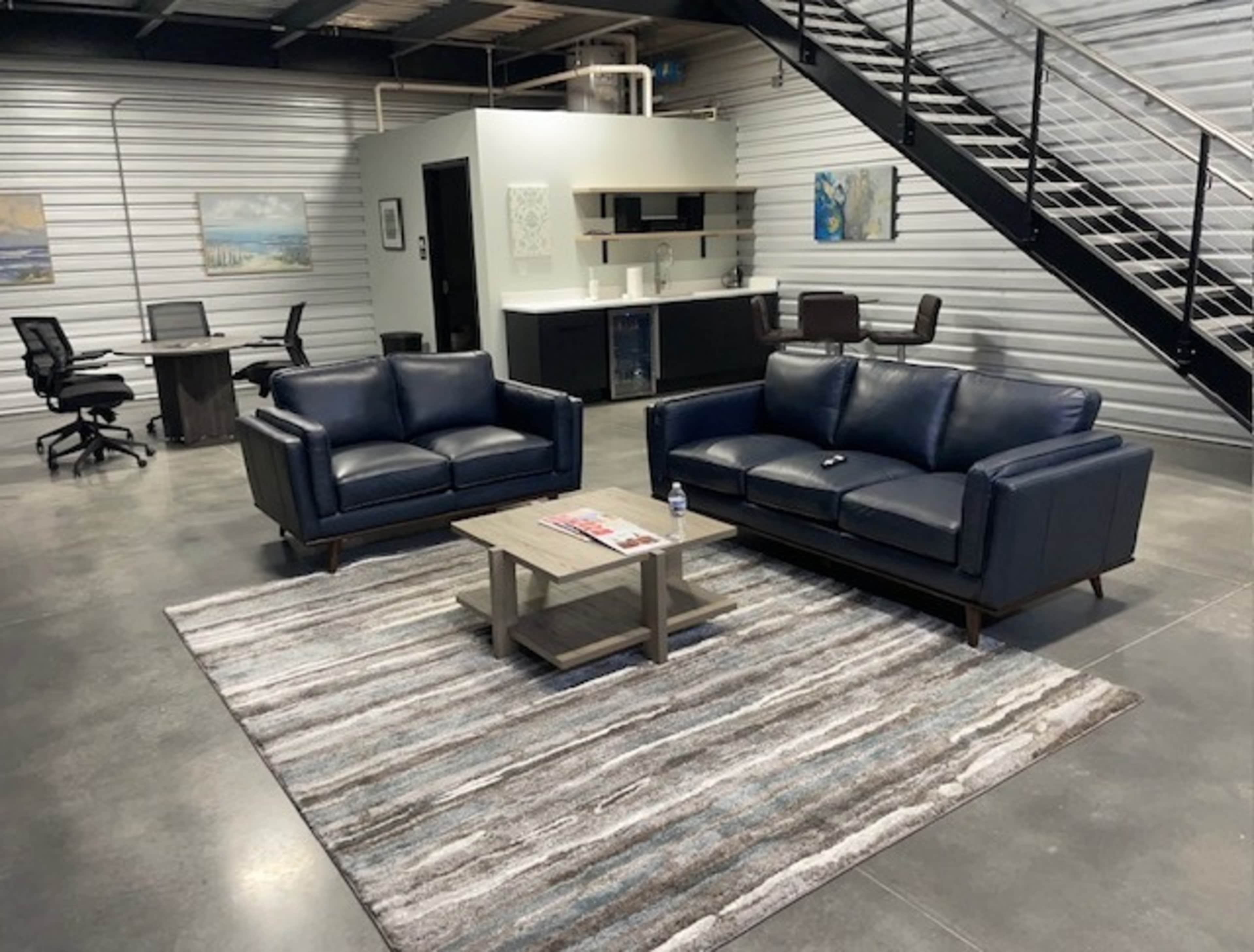 The image shows a modern, industrial-style office space featuring two blue sofas, a coffee table, and a kitchen area adjacent to a staircase.