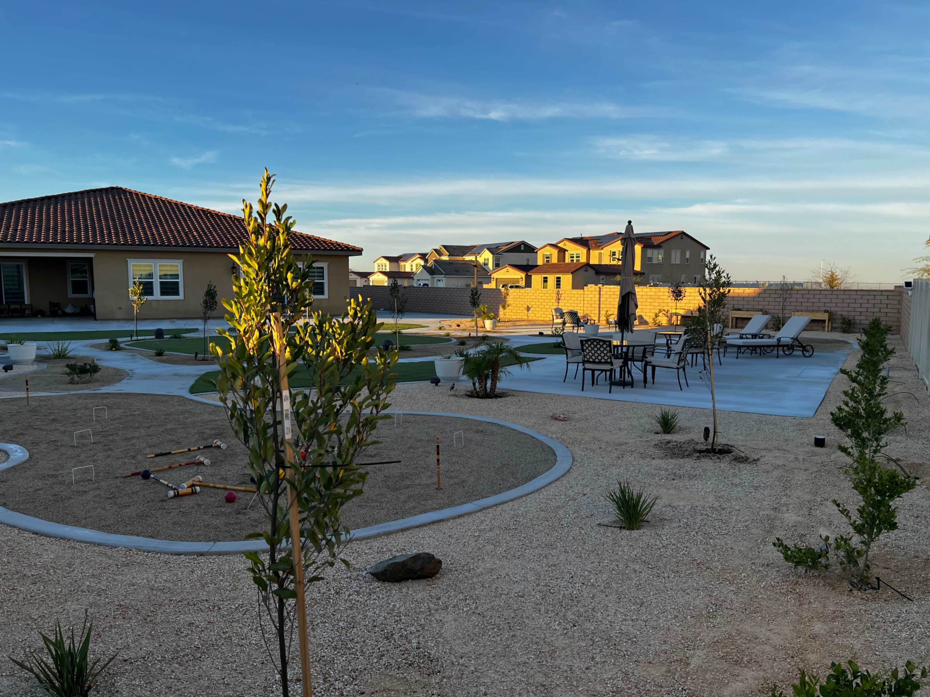 The image shows a landscaped backyard with a gravel surface, seating area, and a few trees under a clear sky.