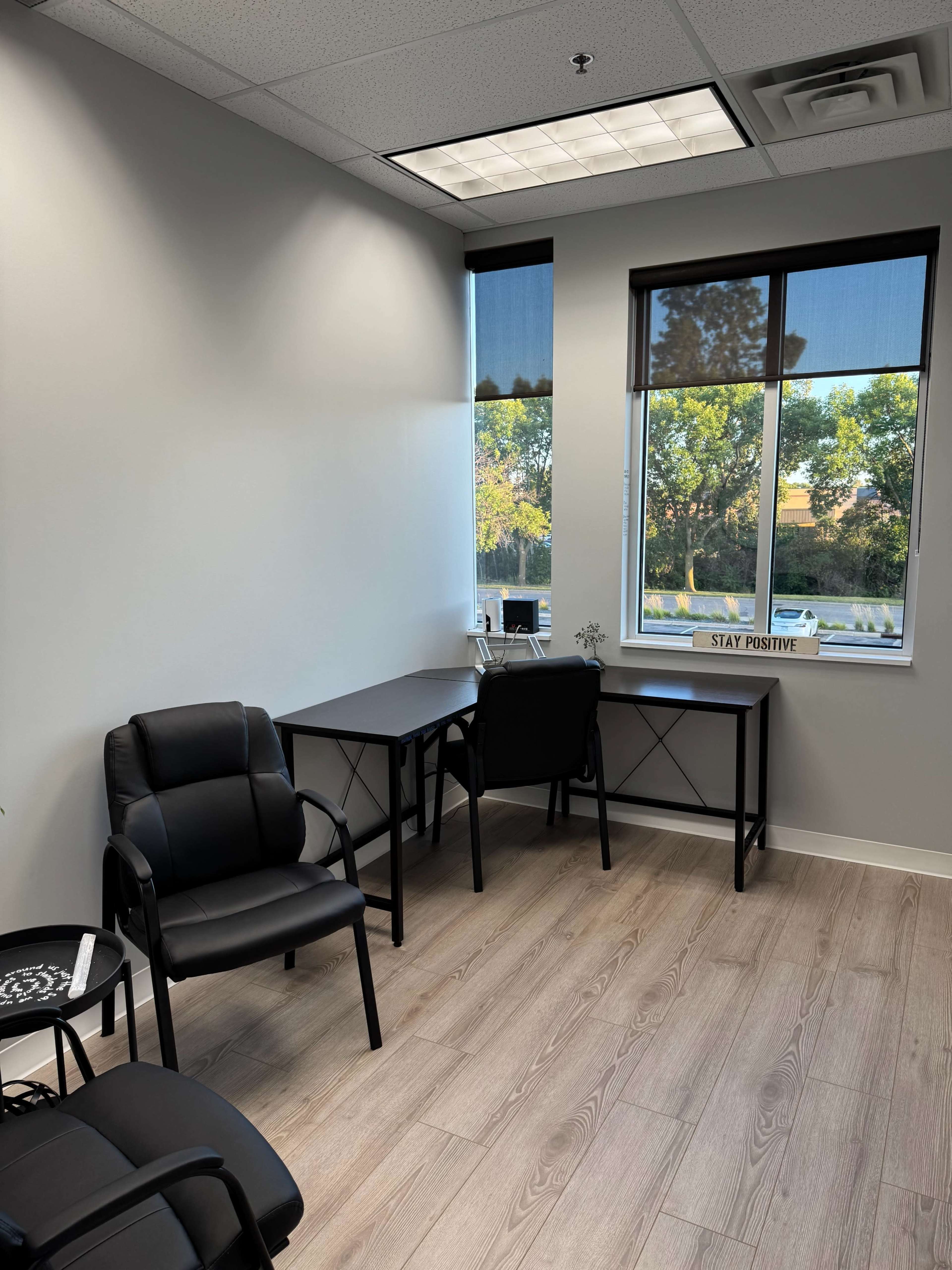 The image shows a minimalistic office space with two black chairs and a desk positioned near a window that overlooks a green landscape.