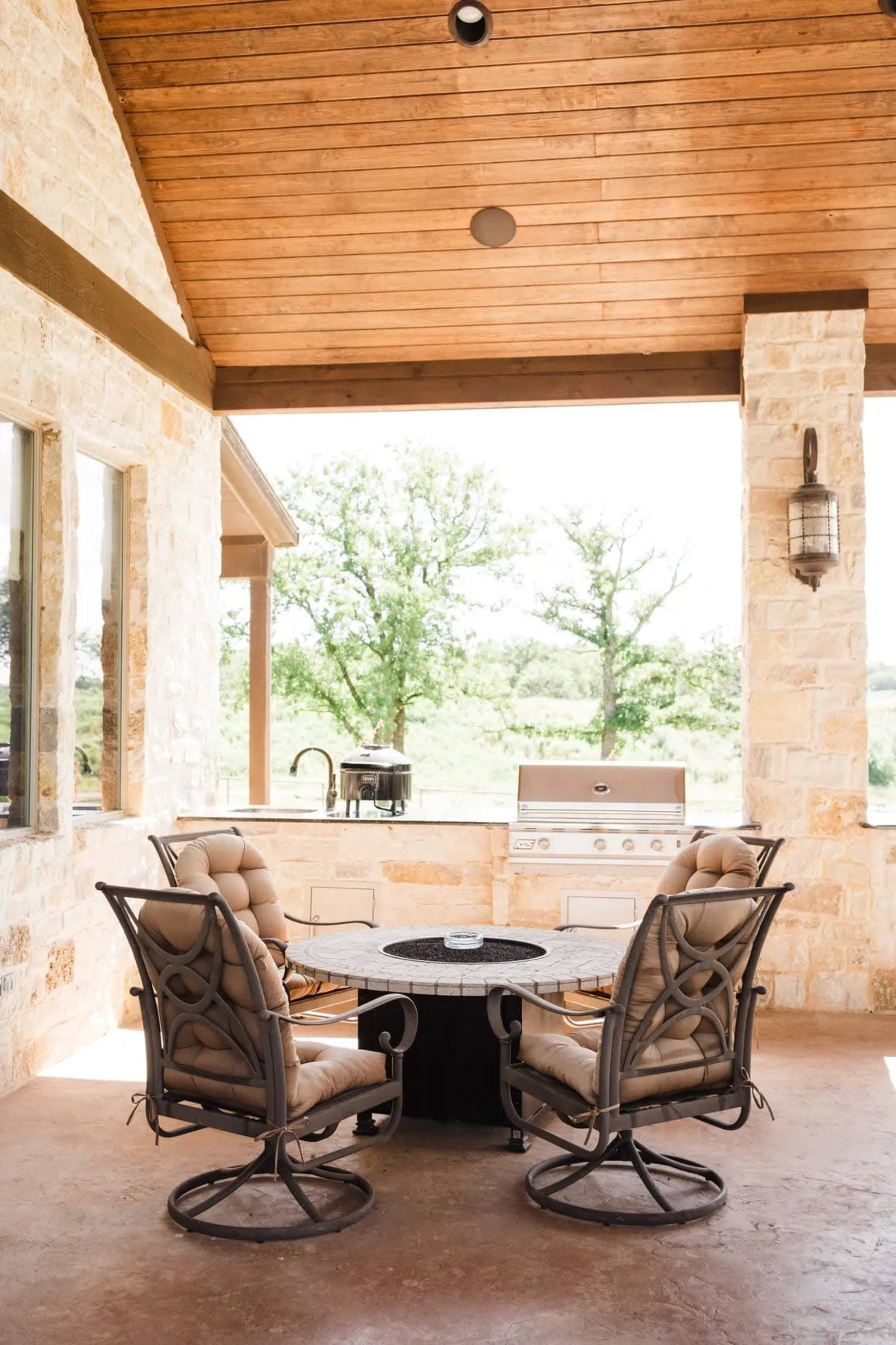 The image shows a patio area with two cushioned chairs around a circular table, featuring an outdoor kitchen with a grill in the background and trees visible through large windows.