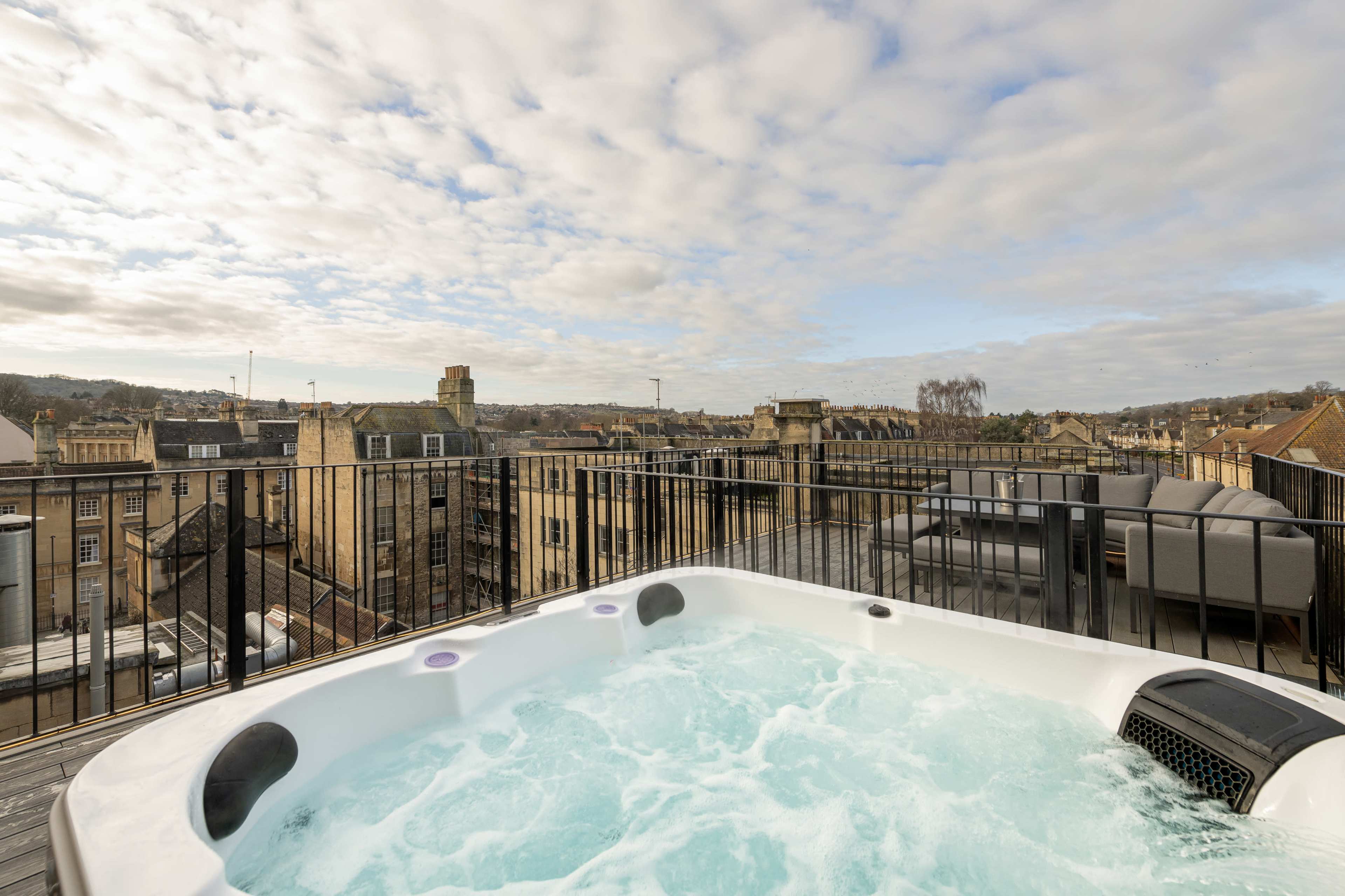 A rooftop hot tub is situated on a balcony overlooking a cityscape with buildings under a cloudy sky.