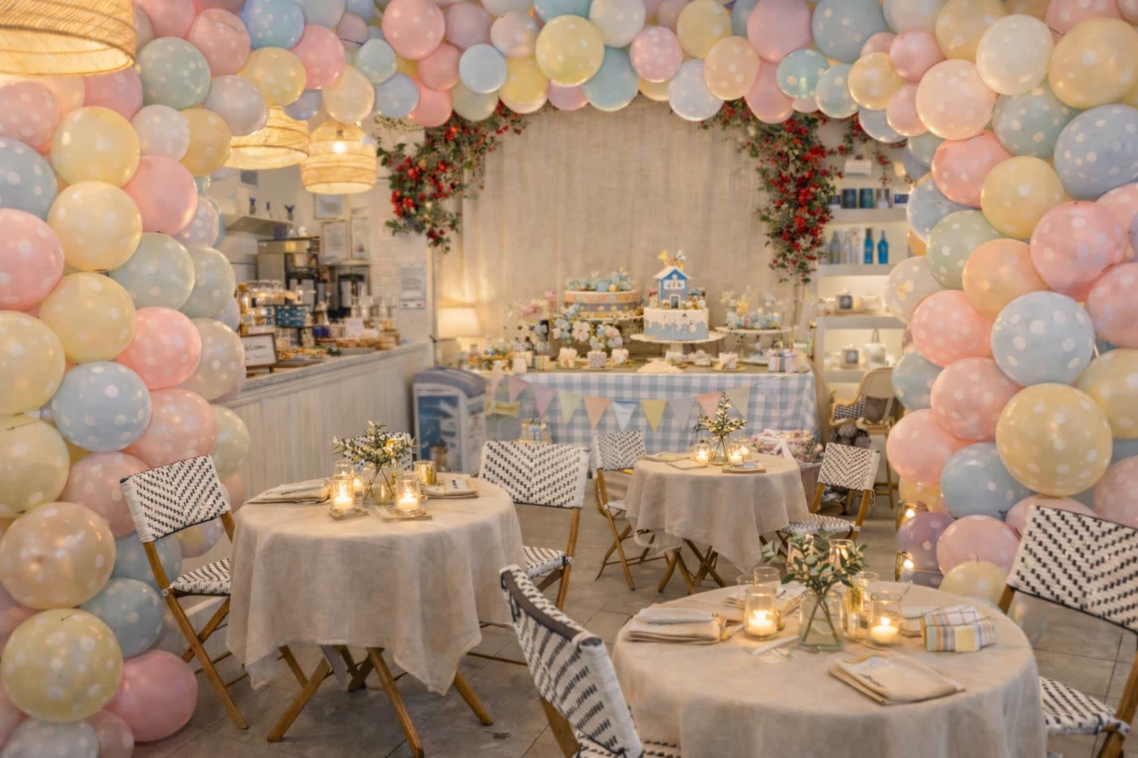 A festive event space features round tables with beige tablecloths, surrounded by a decorative arch made of pastel balloons and adorned with flowers, with a dessert display in the background.