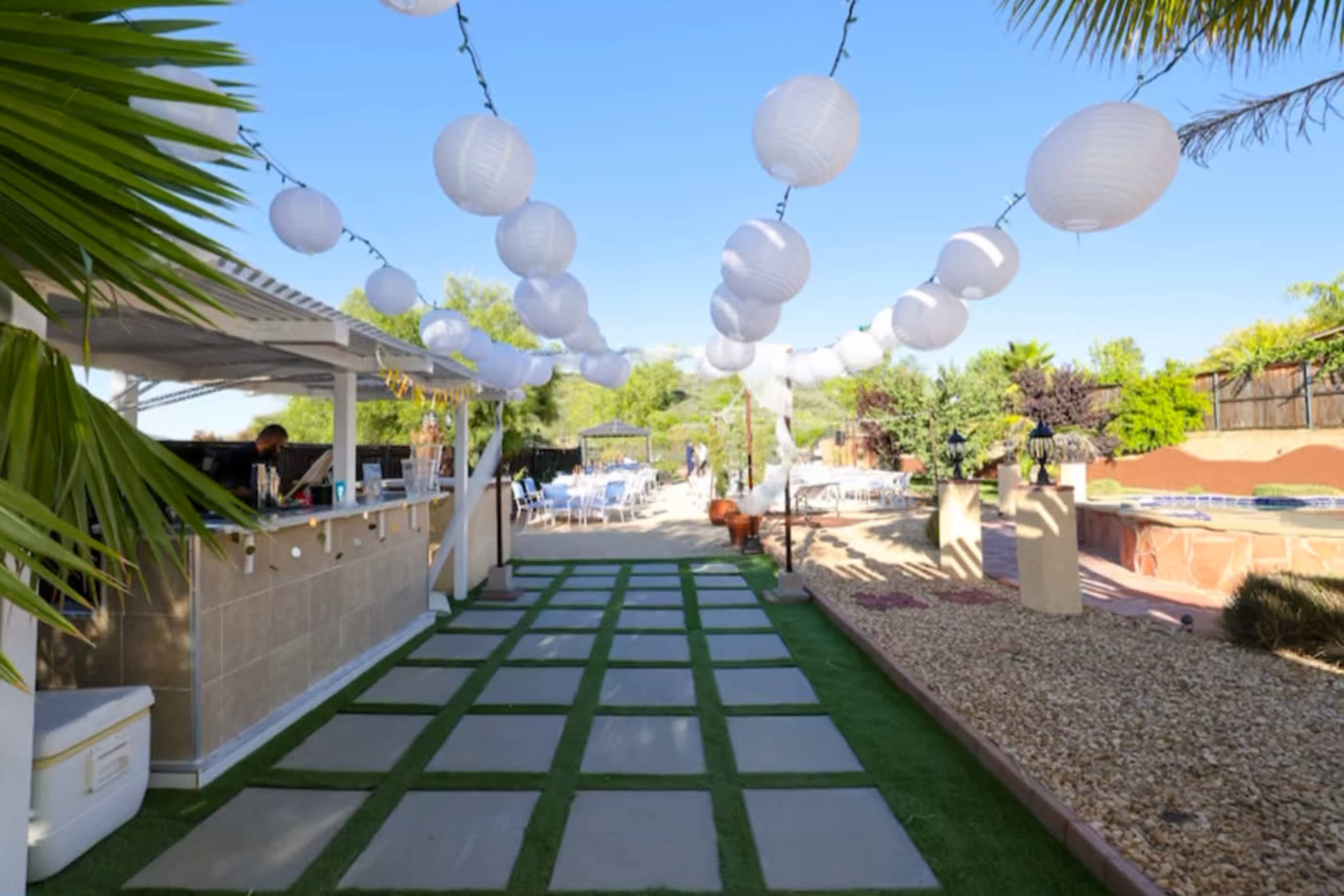 A pathway lined with stone tiles leads through a garden area adorned with hanging lanterns and a bar on the left.