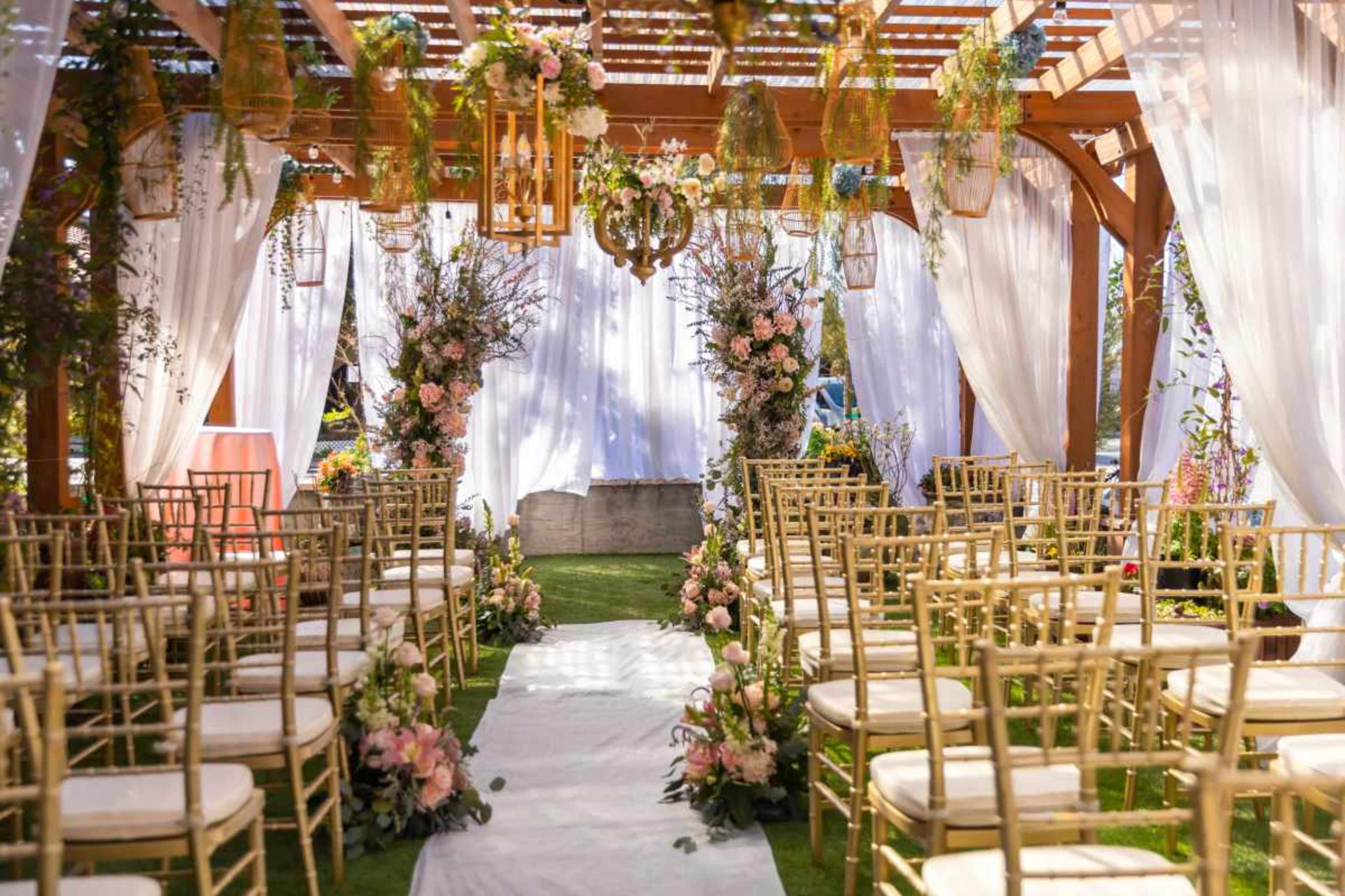 The image shows a wedding ceremony setup with rows of gold chairs facing a floral arch and decorative elements under a wooden pergola.