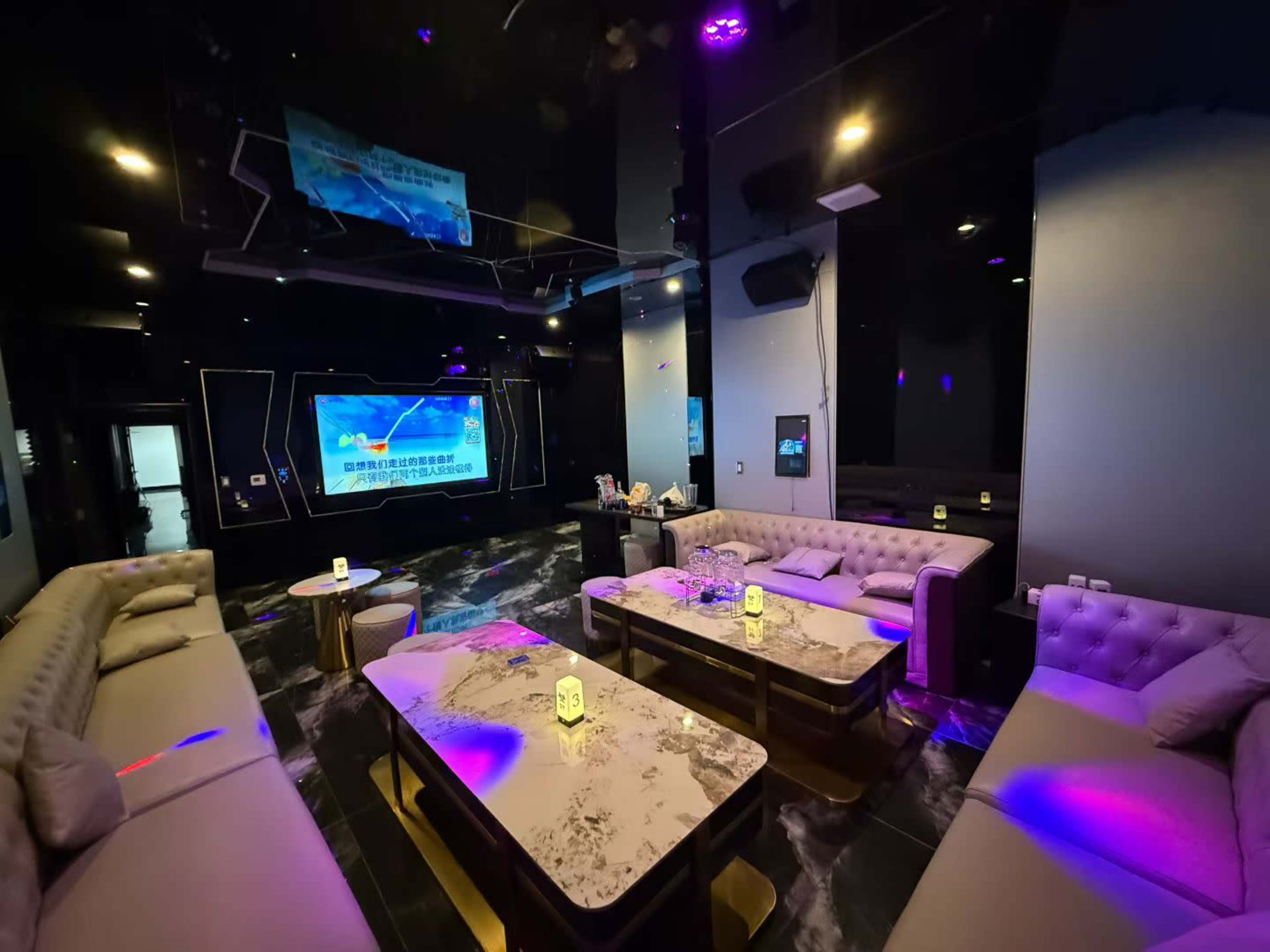 The image shows a modern lounge area with dark decor, featuring large sofas and tables, illuminated by colorful lights and a large screen on the wall.