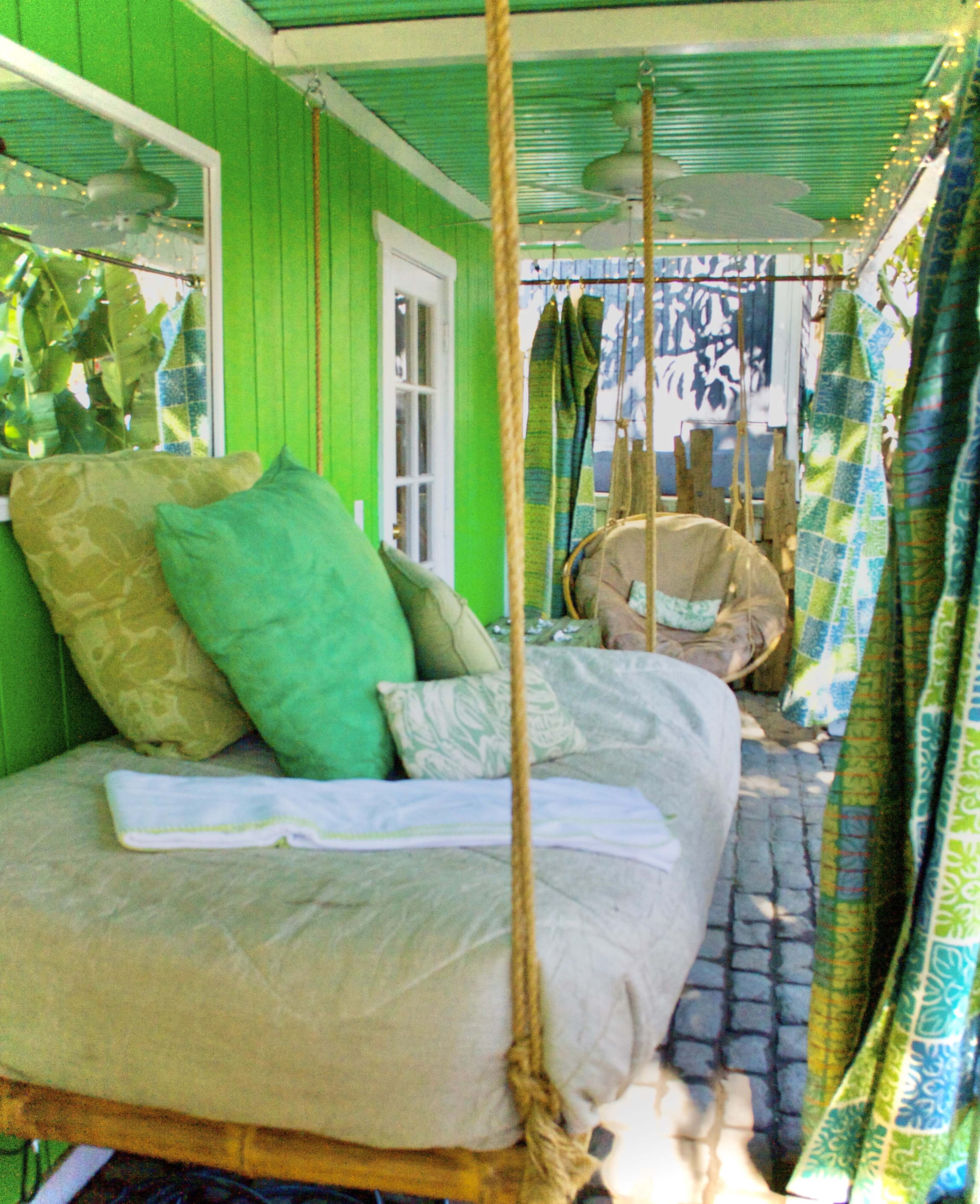 The image shows a brightly painted green porch with a hanging bed and a cozy, woven chair surrounded by decorative fabrics.