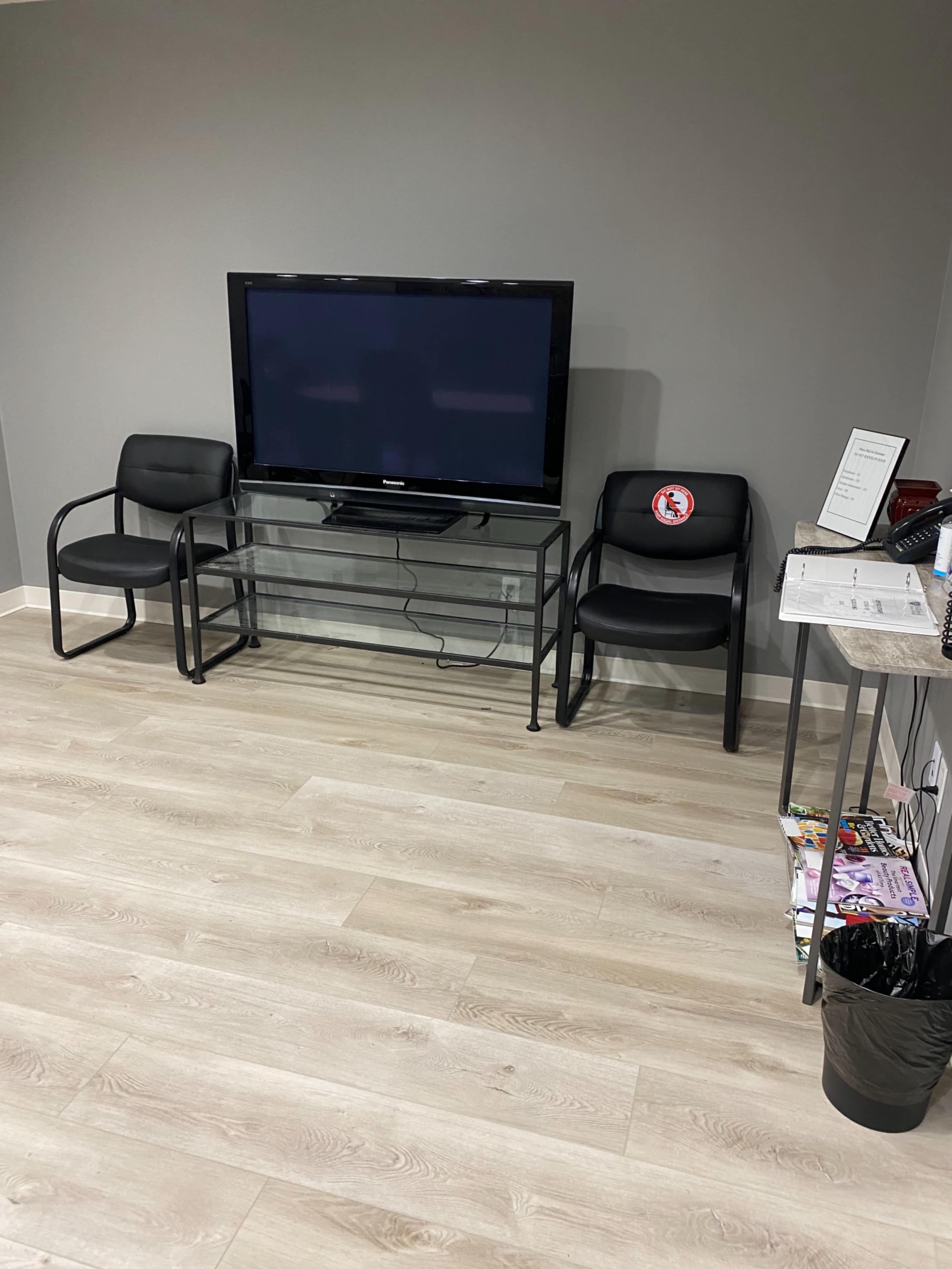 A waiting area with two black chairs beside a glass TV stand, which holds a television against a gray wall, with a magazine rack and a trash bin nearby.