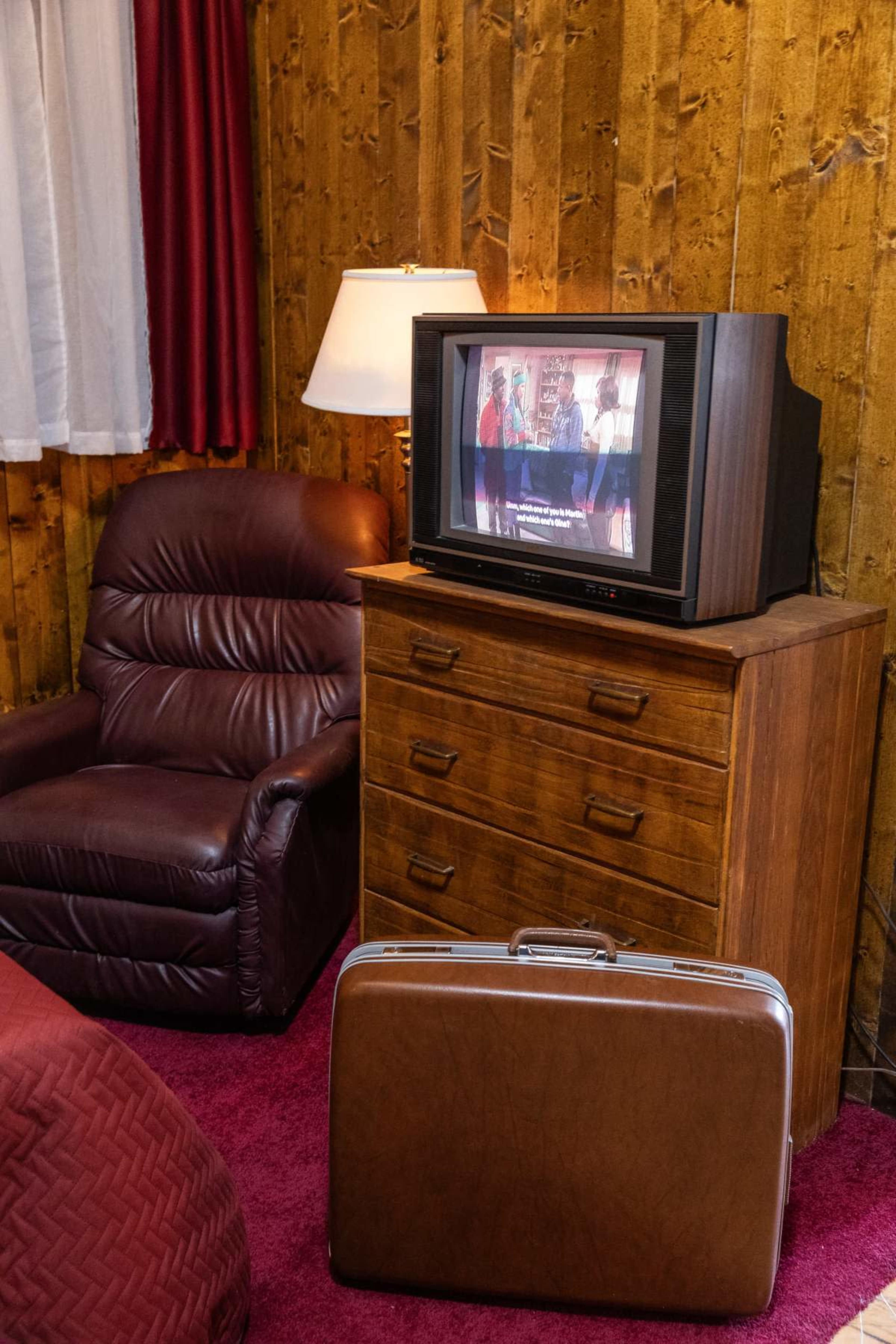 The image features a wooden dresser with a vintage television on top, a recliner next to it, and a suitcase on the carpeted floor in a room with wooden walls.