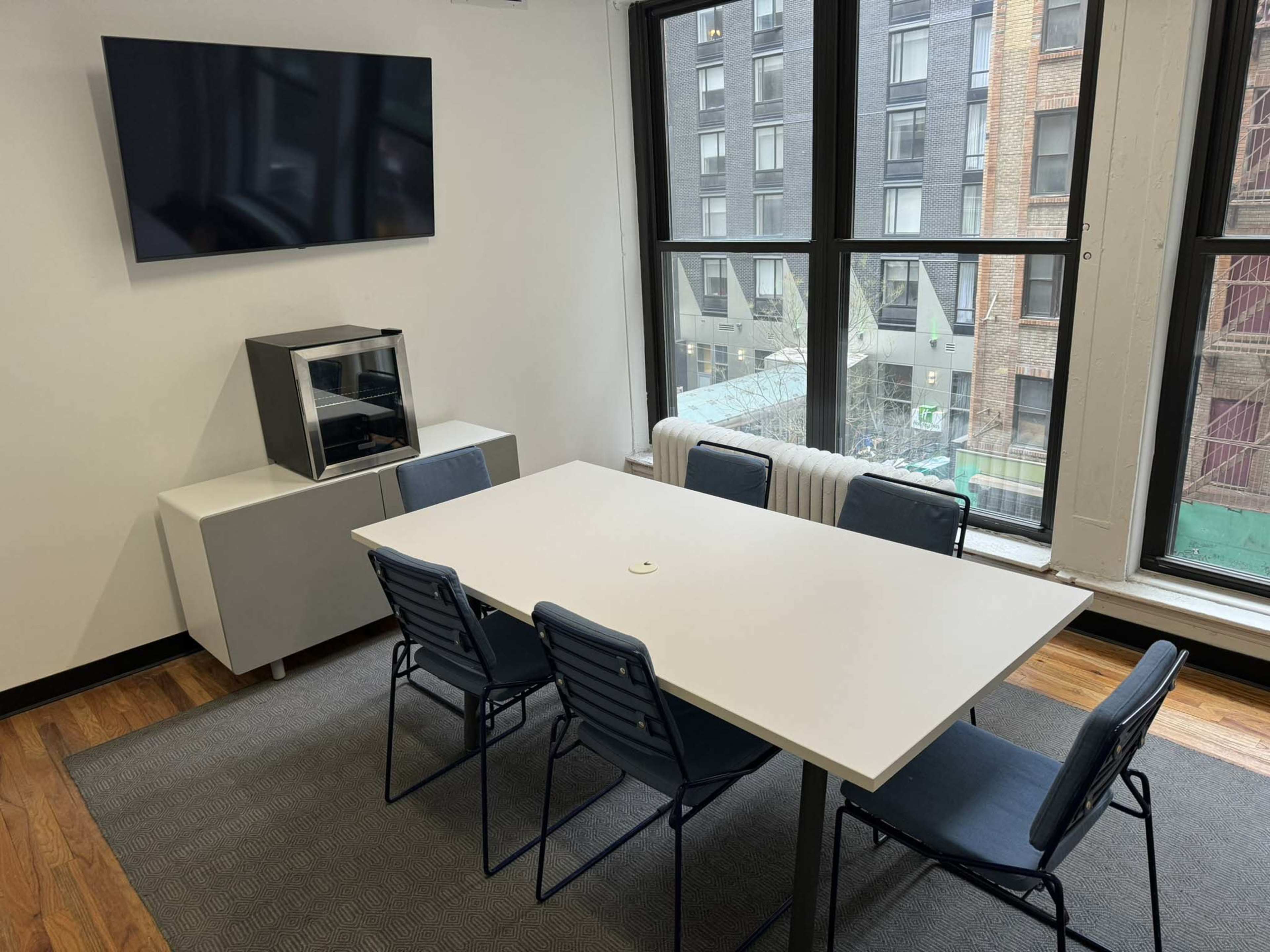 A conference room features a white table surrounded by six chairs, with a television mounted on the wall and large windows offering a view of an adjacent building.