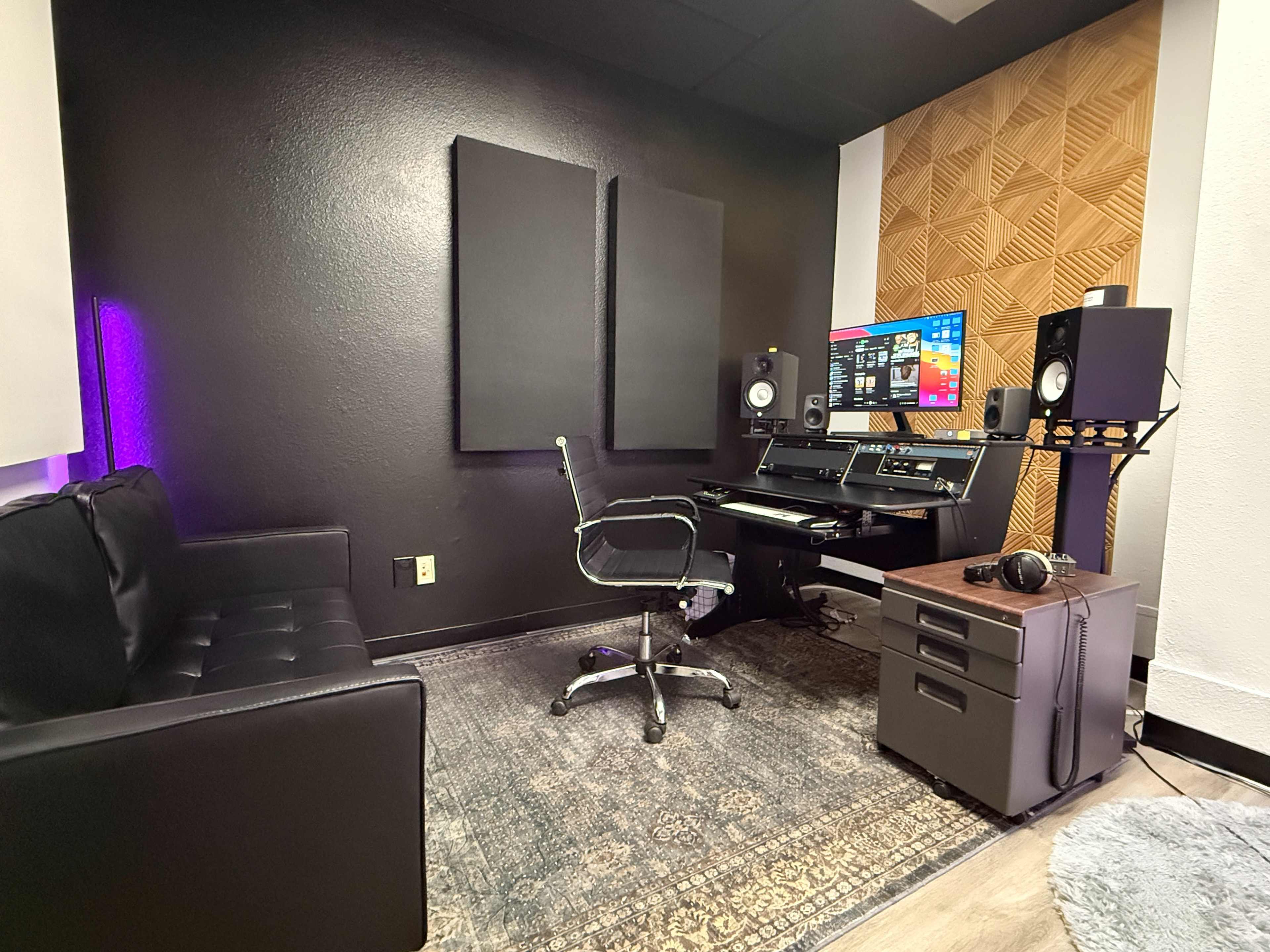 The image shows a modern music studio featuring a desk with a computer setup, a swivel chair, a couch, and acoustic panels on the walls.