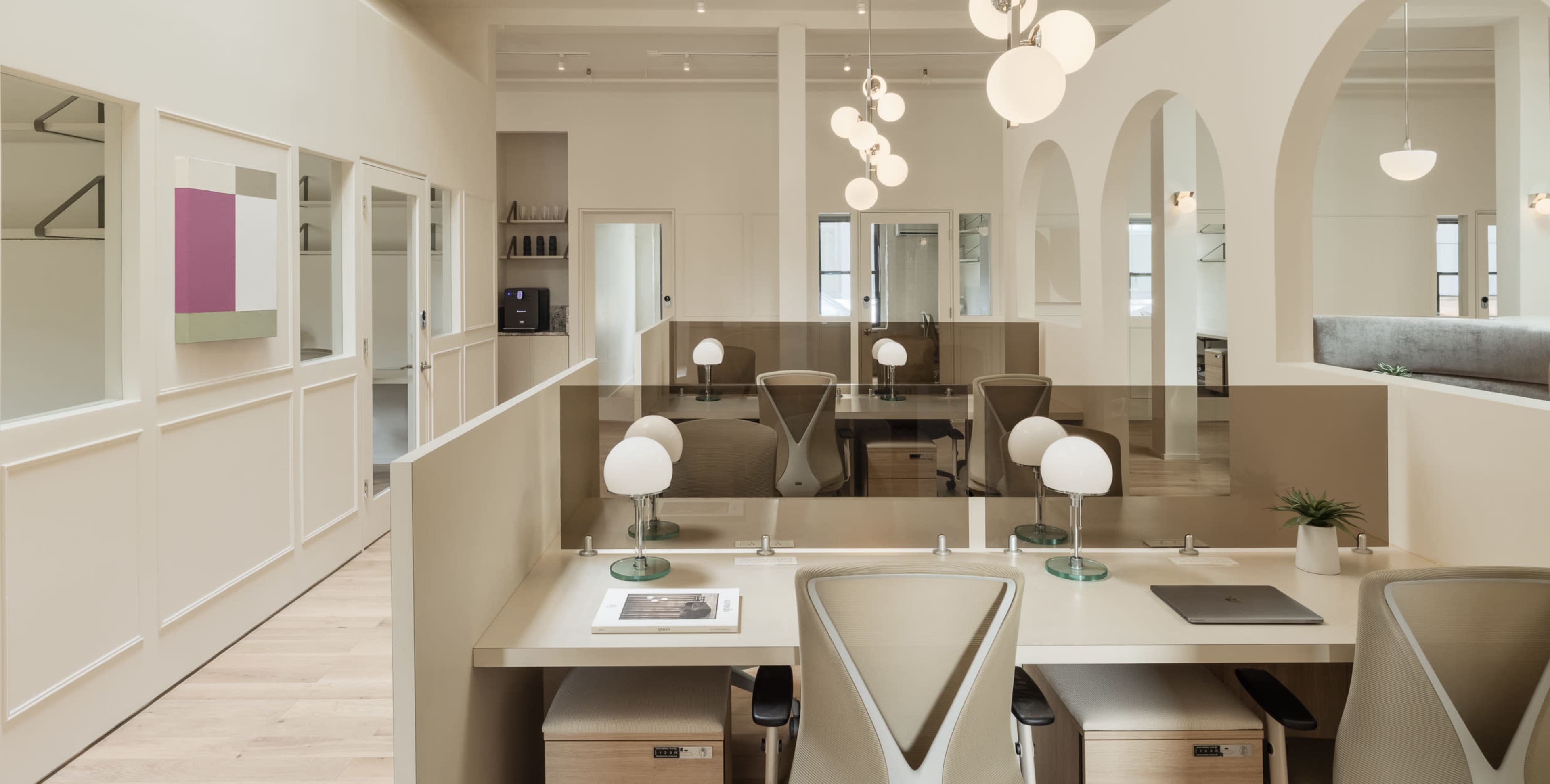 A modern office space with light-colored walls, arched doorways, and desks equipped with lamps and partitions.