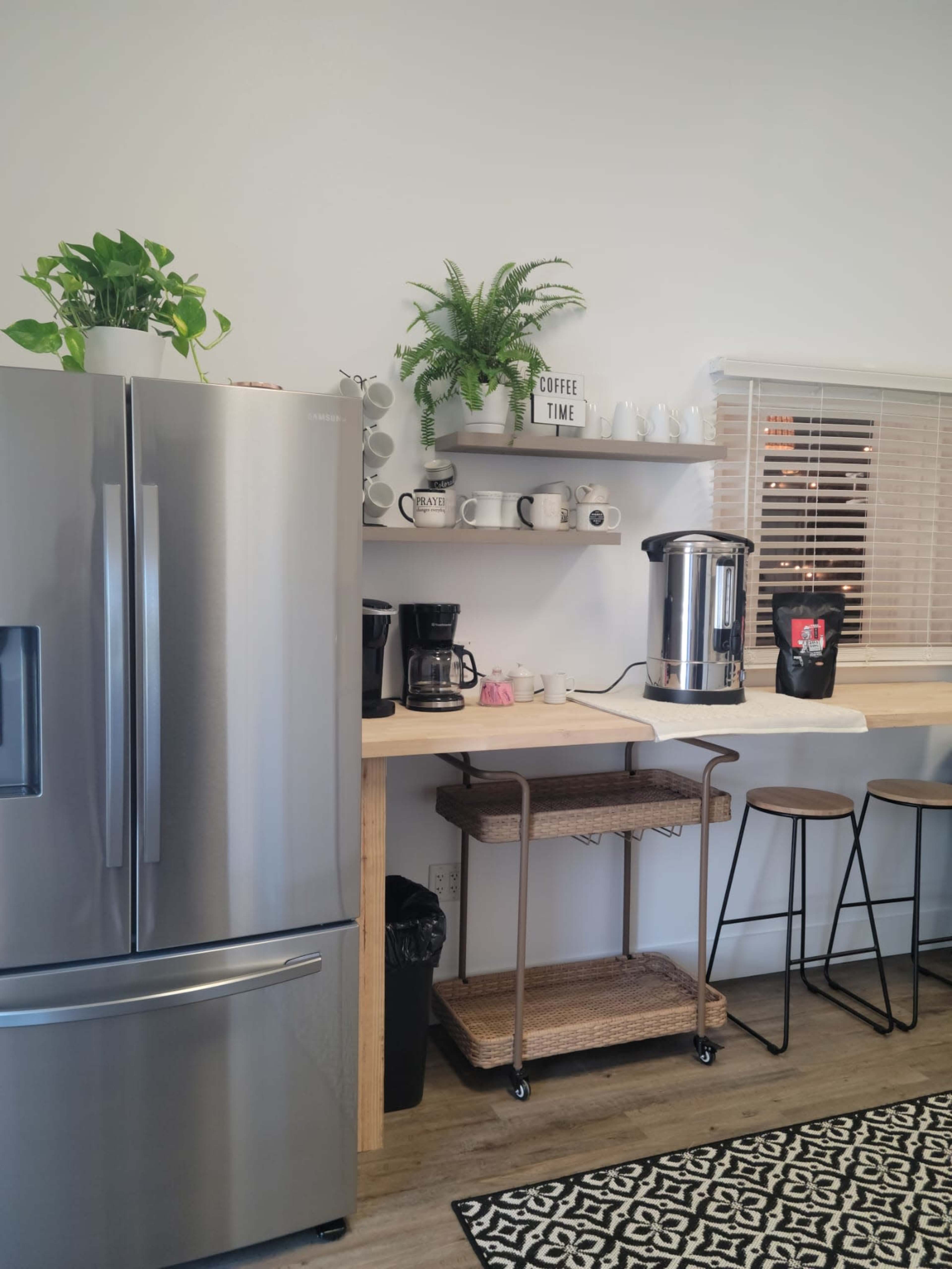 A modern kitchen features a stainless steel refrigerator, a coffee station with multiple coffee makers, and a bar with stools, all arranged against a bright, minimalistic backdrop.