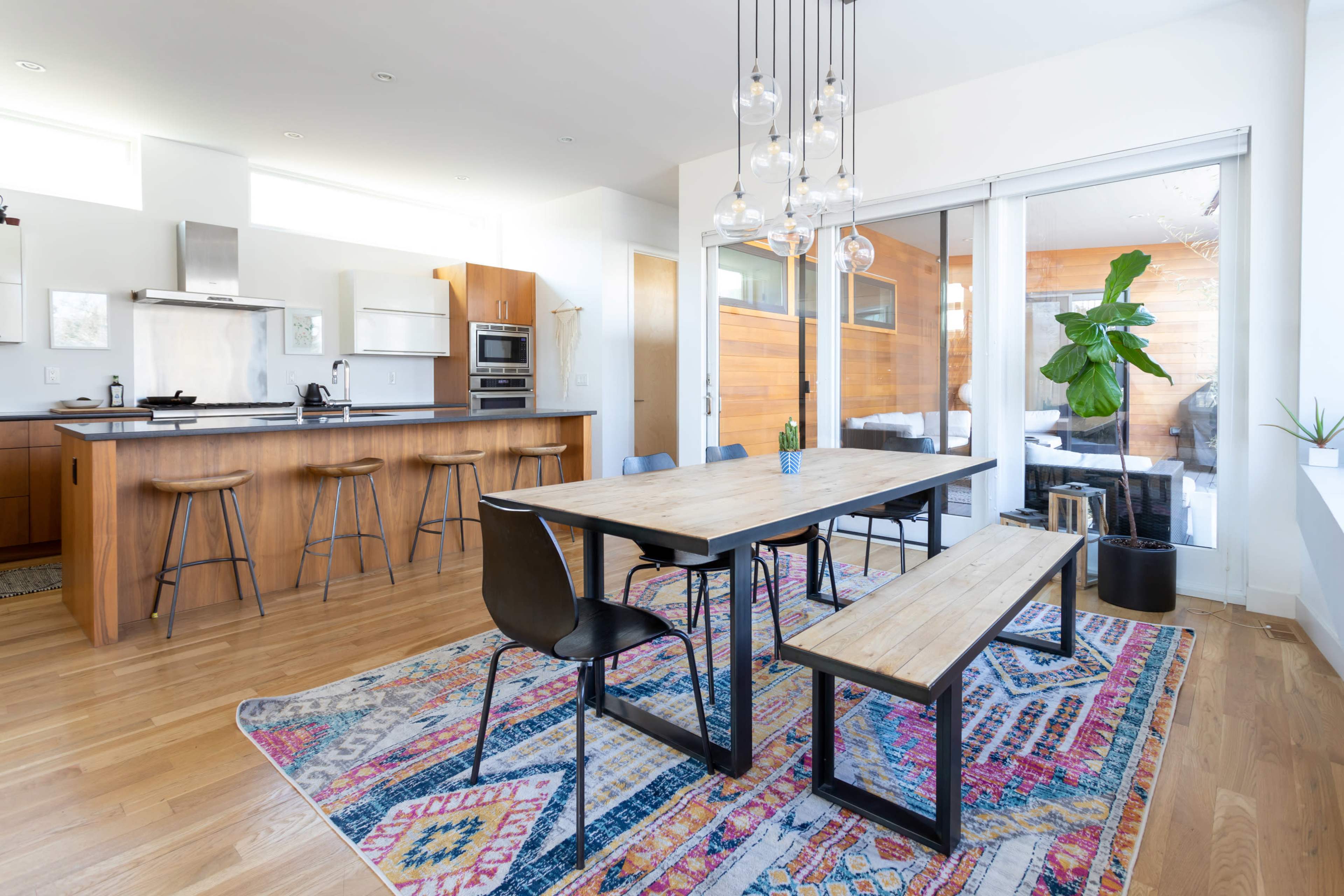 A modern kitchen and dining area with a wooden table surrounded by chairs, bar stools at the kitchen counter, and large windows allowing natural light to enter.