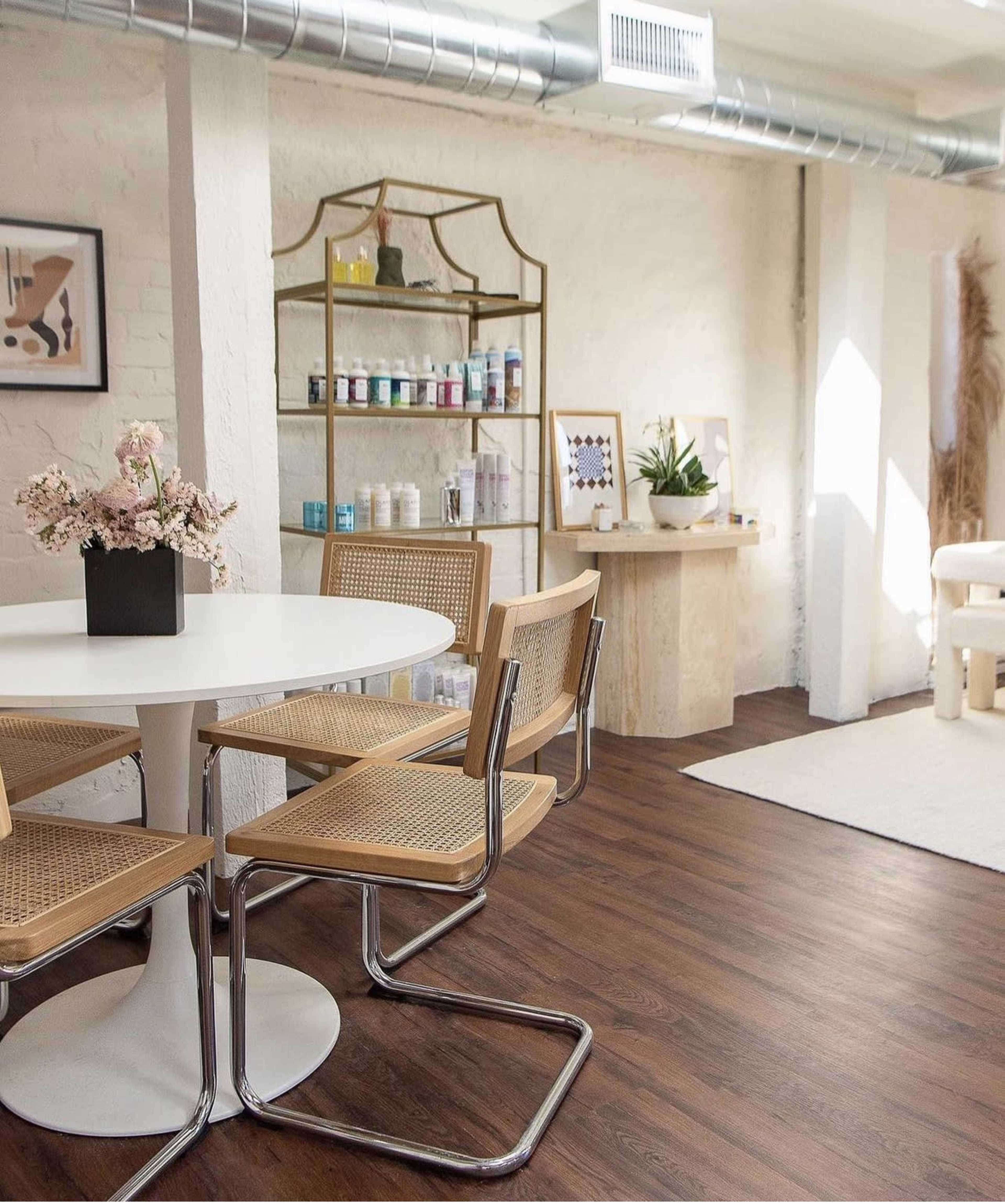 A bright, modern interior with a round white table surrounded by four cane-back chairs, alongside a decorative shelf displaying various bottles and plants.
