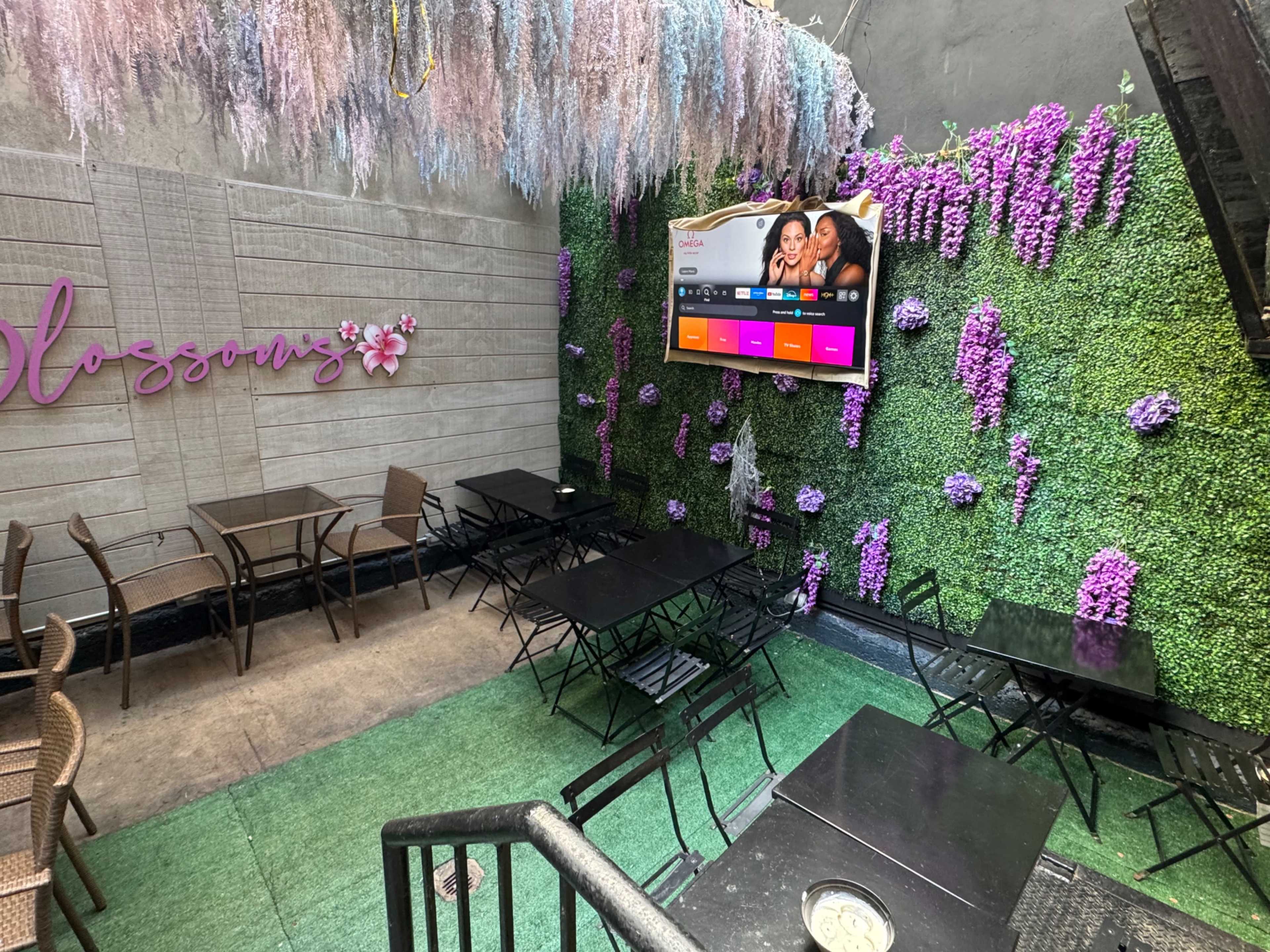 The image shows a cozy outdoor seating area with black tables and chairs, surrounded by a wall covered in purple flowers and greenery, featuring a screen displaying colorful content.