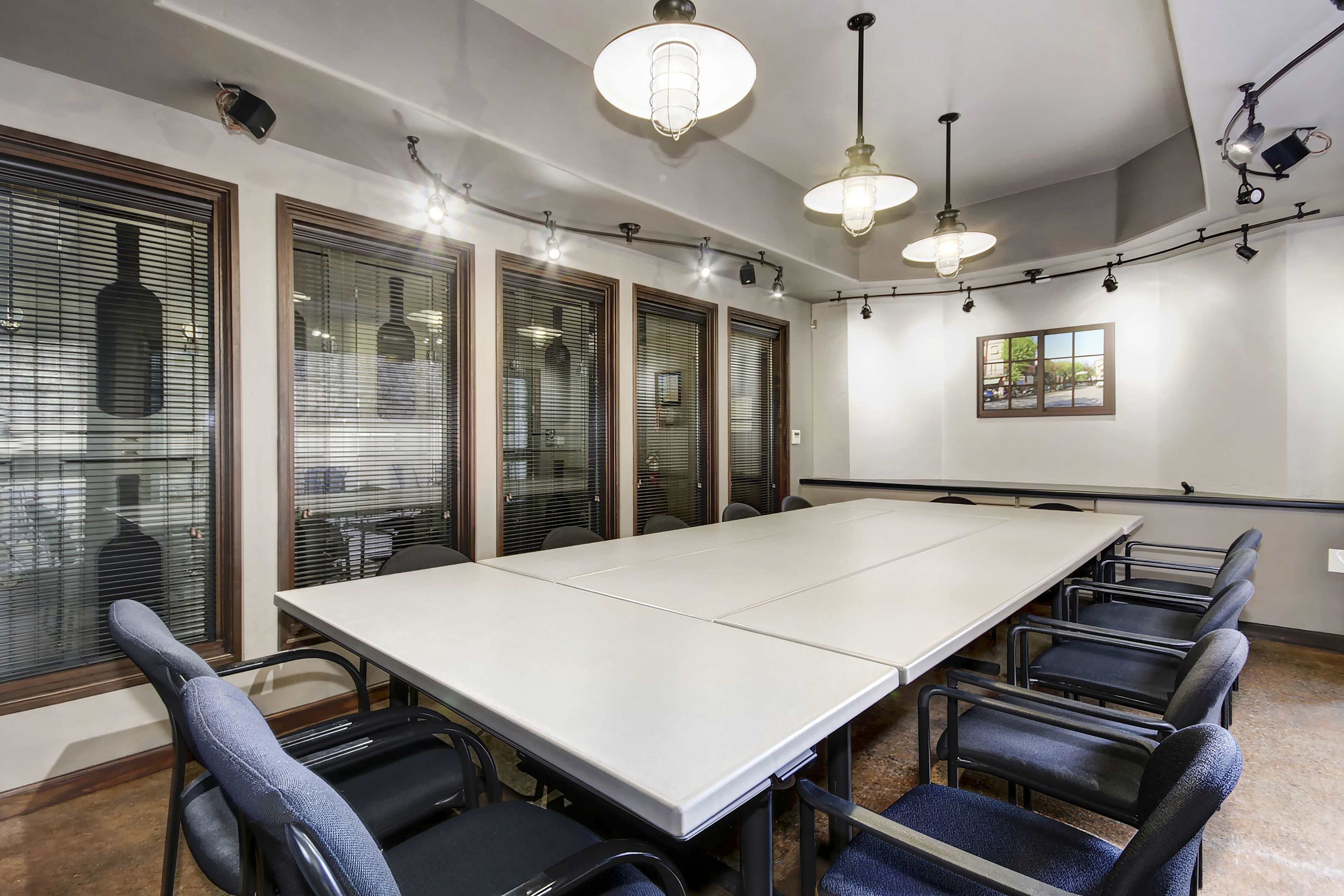 A conference room features a large rectangular table surrounded by black and blue chairs, with windows on one side and pendant lighting above.