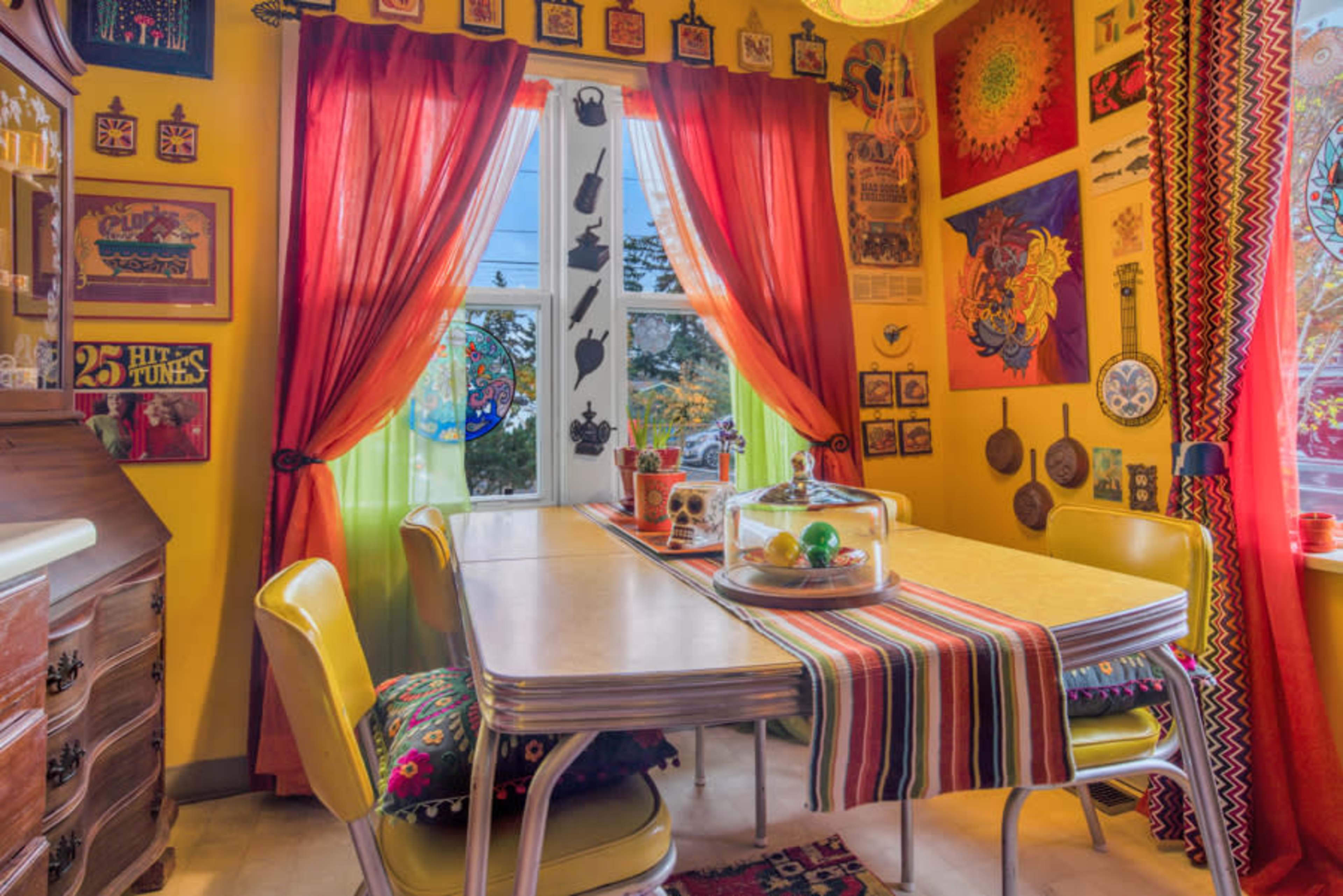 A brightly colored dining area features a table with a striped table runner, surrounded by vintage-style chairs and adorned with various decorative items on the walls.