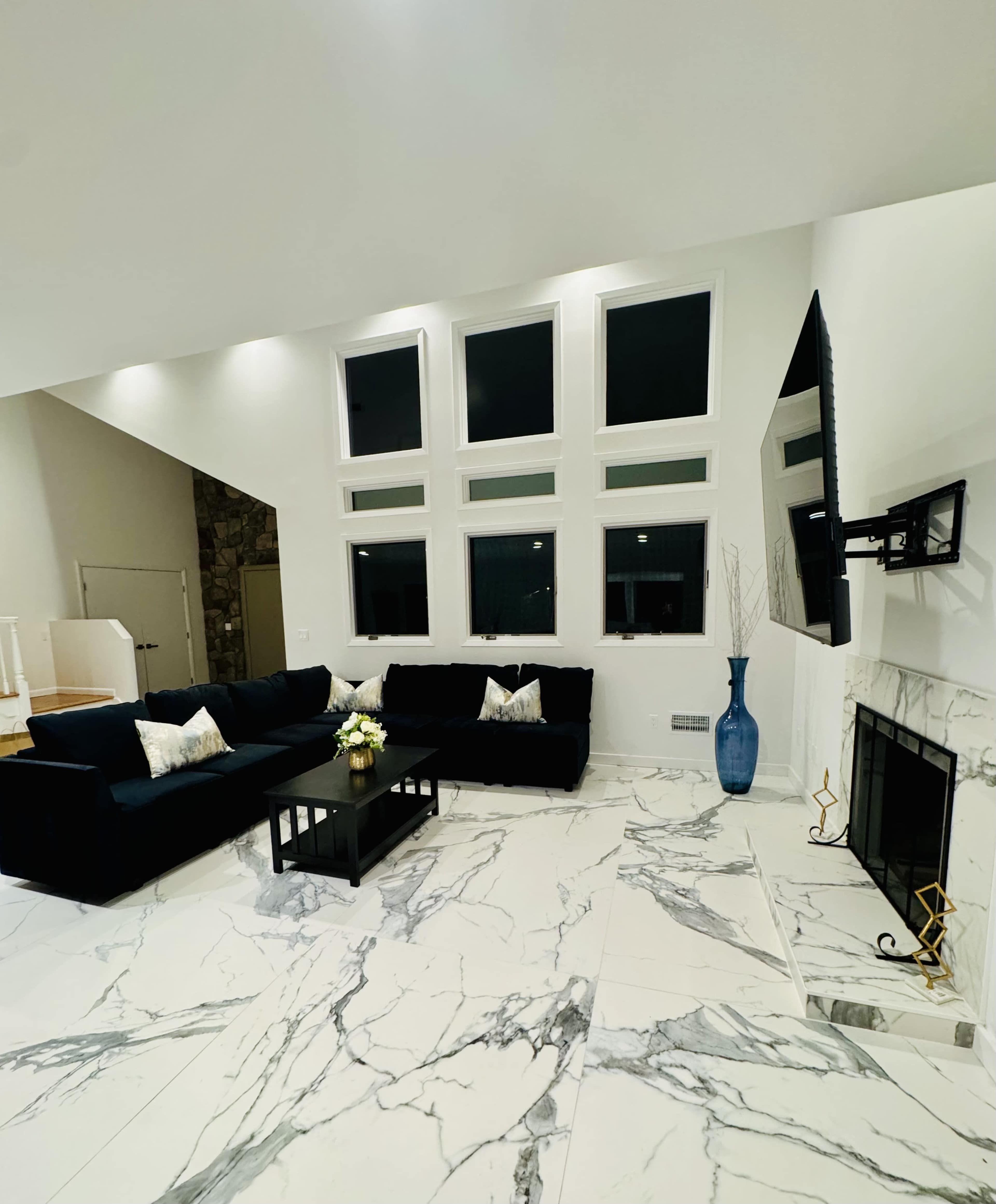 The living room features a large black sectional sofa, a coffee table, and a marble floor, with tall windows allowing natural light to enter the space.