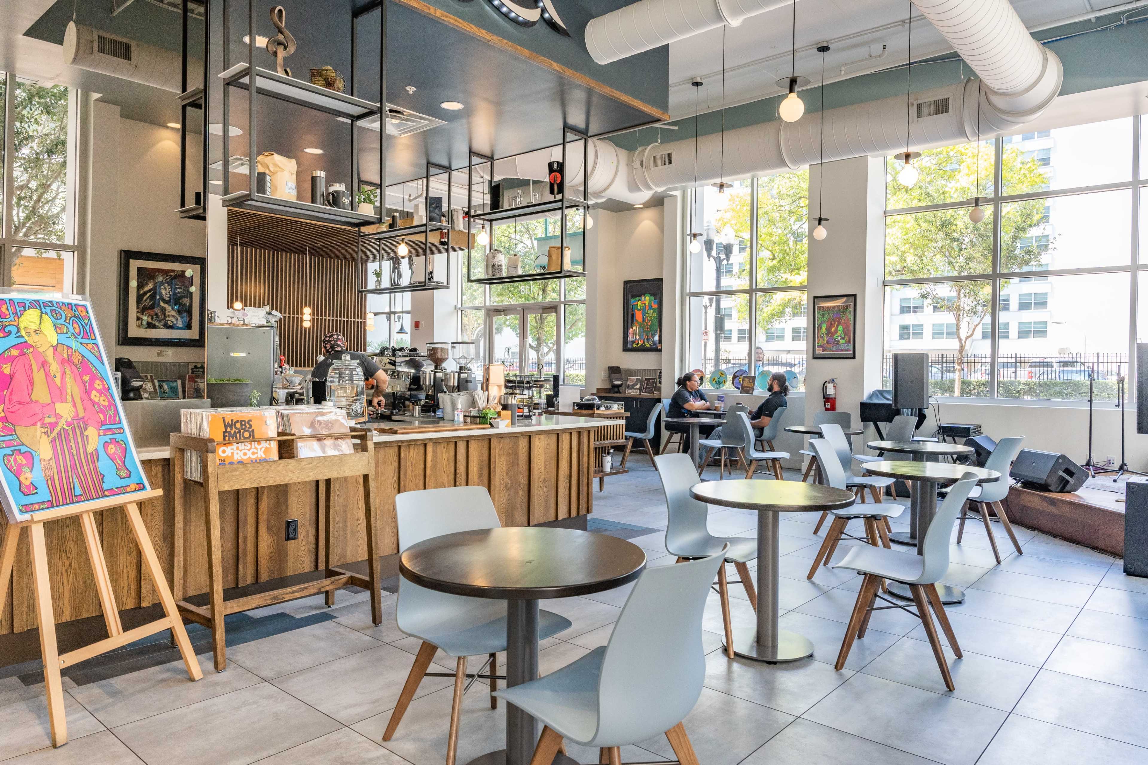 The image shows a brightly lit café with wooden furniture, large windows, and a coffee bar displaying various beverages and artwork on the walls.