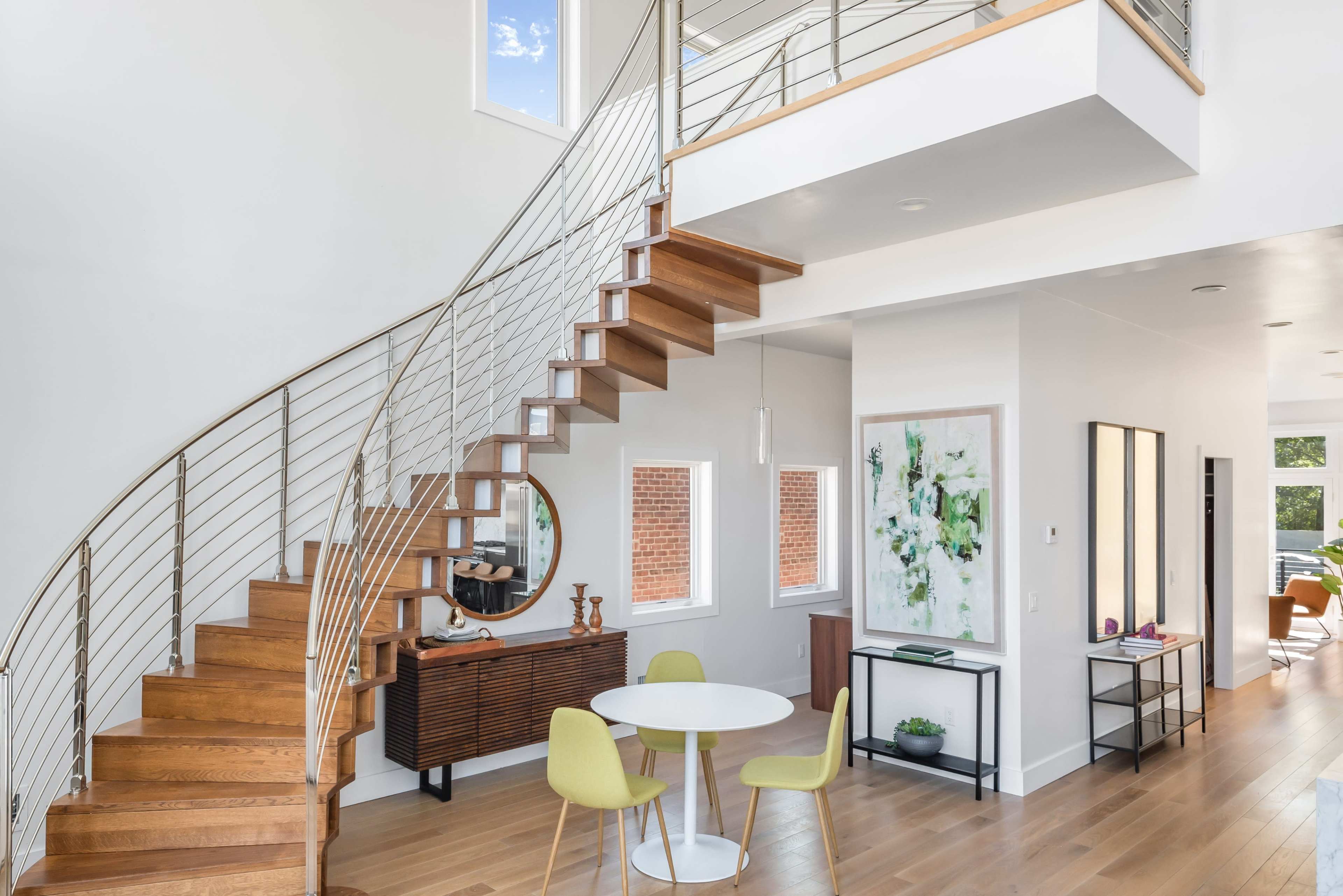 A modern interior featuring a spiral wooden staircase, a round dining table with yellow chairs, and large windows allowing natural light to enter the space.