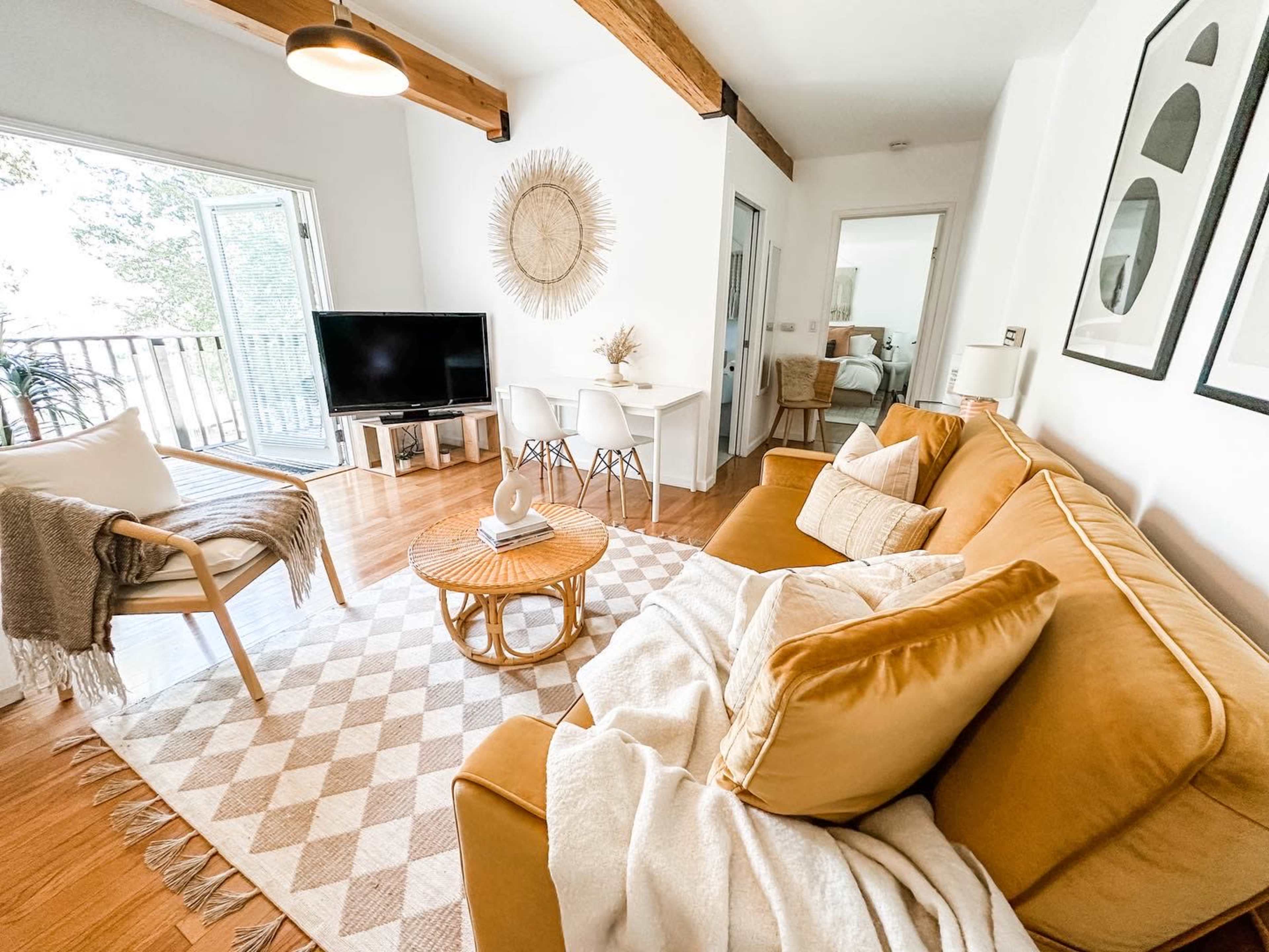 A bright living room features a mustard yellow sofa, a round coffee table, a patterned area rug, and a TV beside large windows.