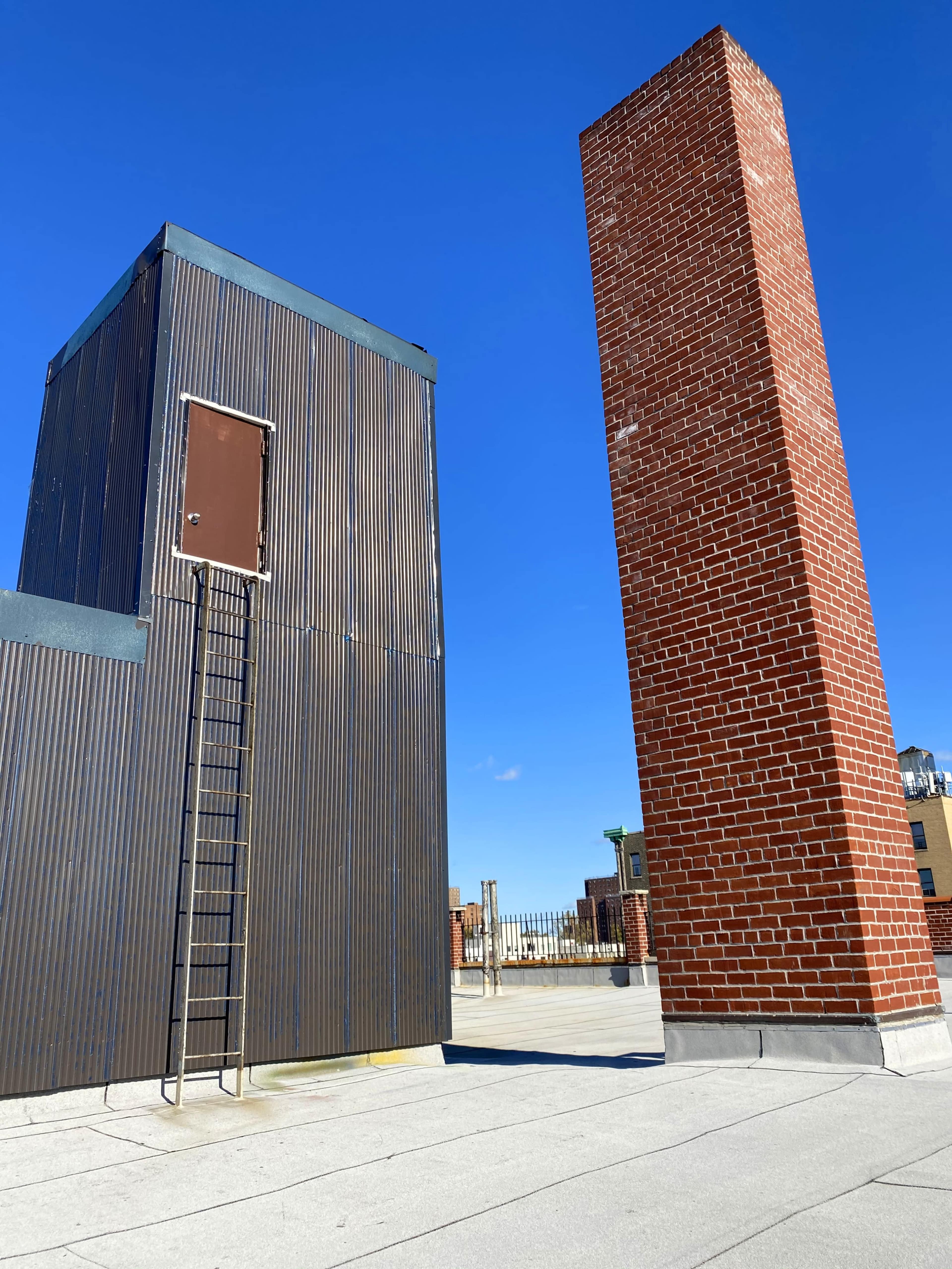 The image shows a rooftop scene featuring a metal structure with a ladder leading to a door, alongside a tall brick chimney under a clear blue sky.