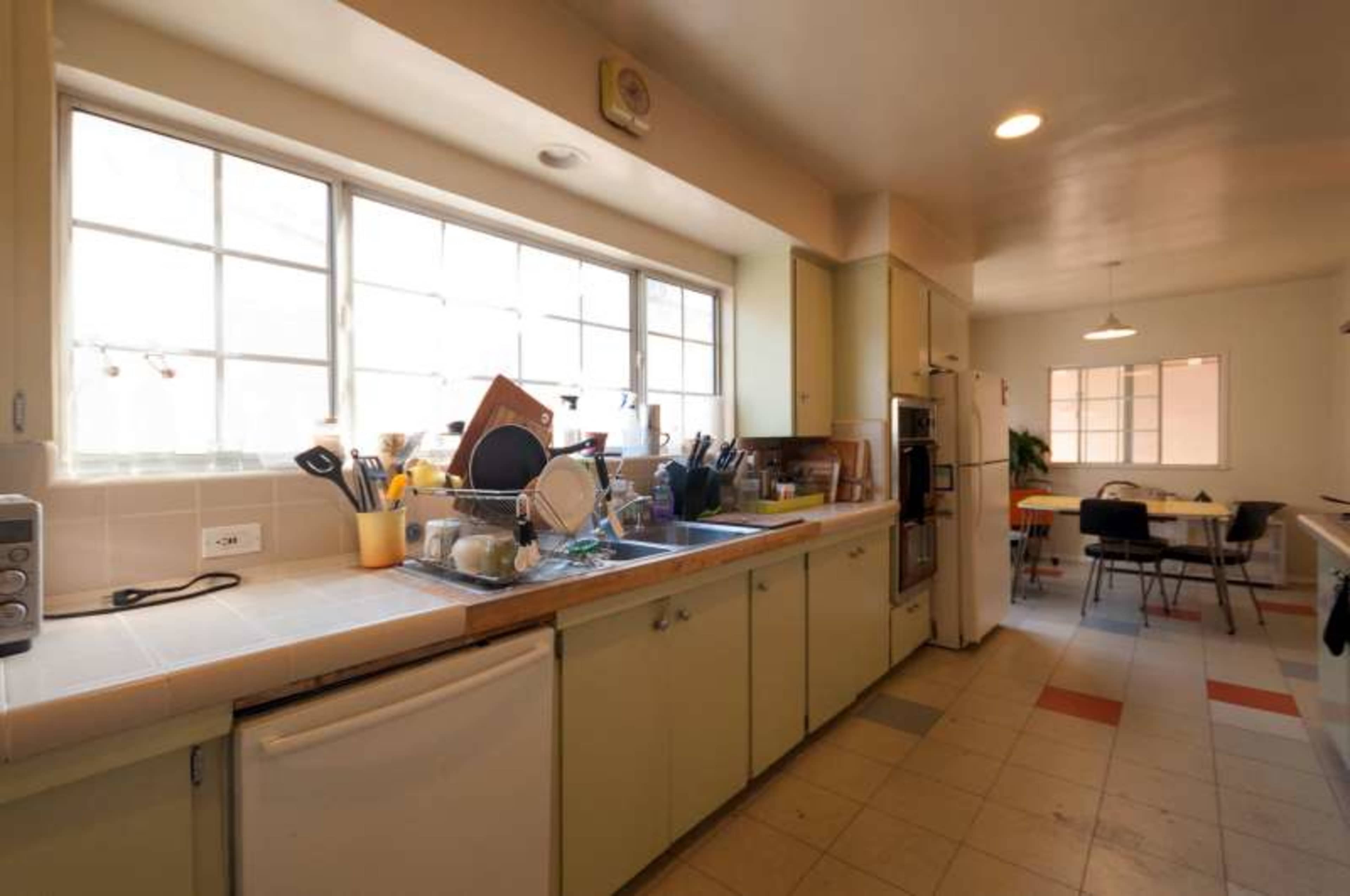 The kitchen features a long countertop lined with dishes, appliances, and utensils, with large windows providing natural light and a dining area visible in the background.