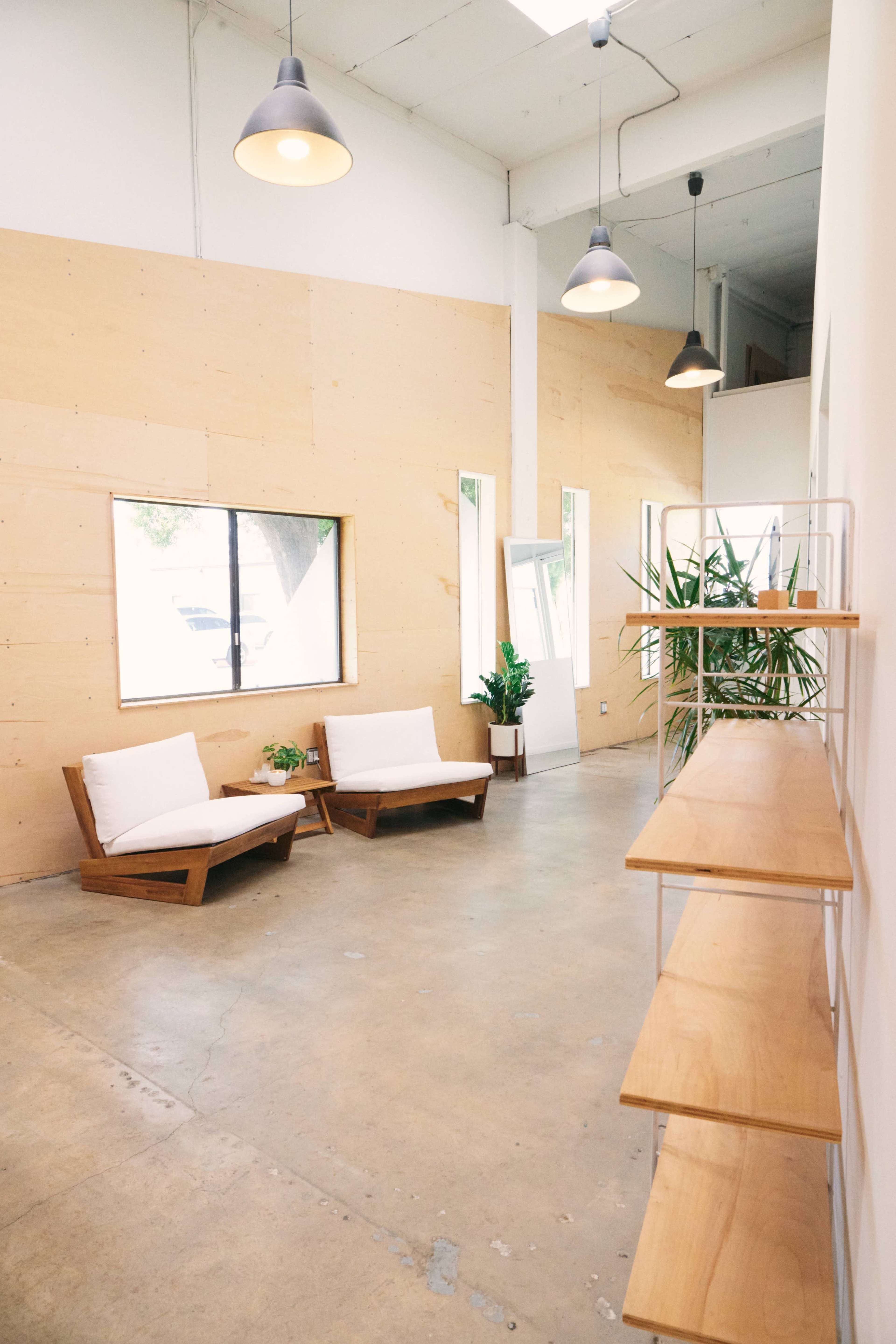 The image shows a modern, minimalist interior with two wooden lounge chairs, a large window, a mirror, potted plants, and a wooden shelf along one wall.
