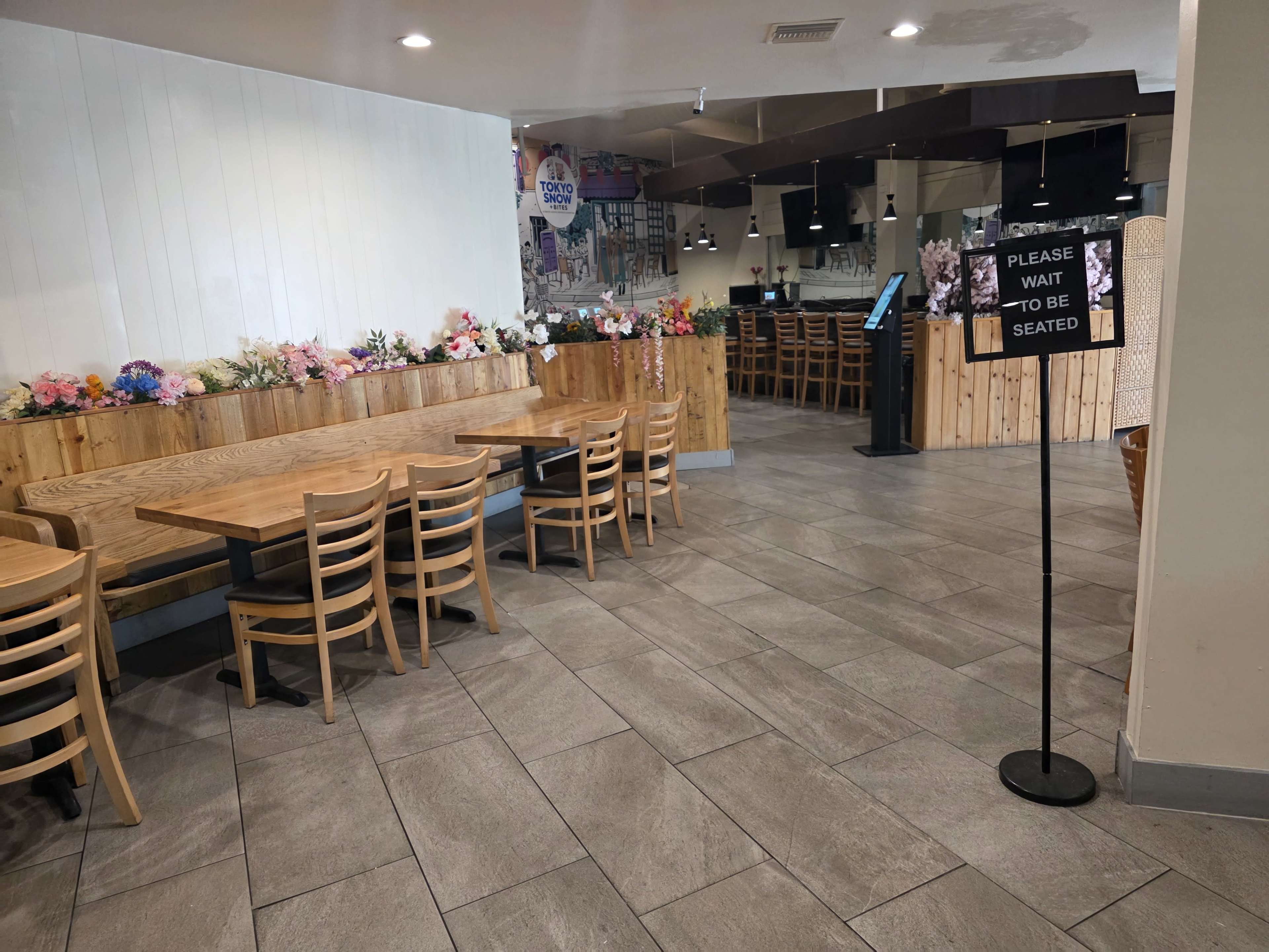 The image shows a restaurant interior with wooden benches and tables arranged in a simple layout, along with a sign indicating that guests should wait to be seated.
