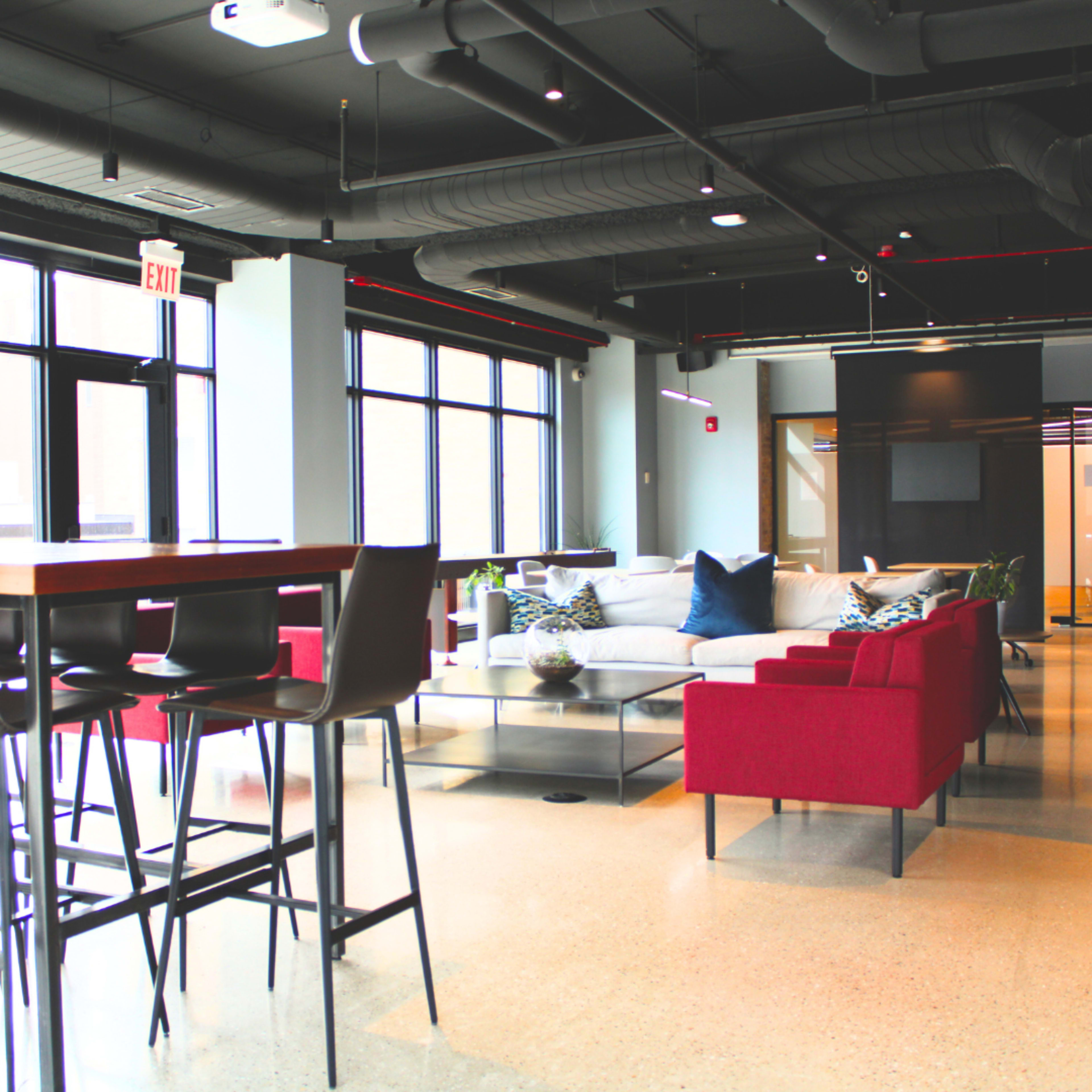 The image shows a modern office lounge area with a sofa, red chairs, a coffee table, and a high table with stools.