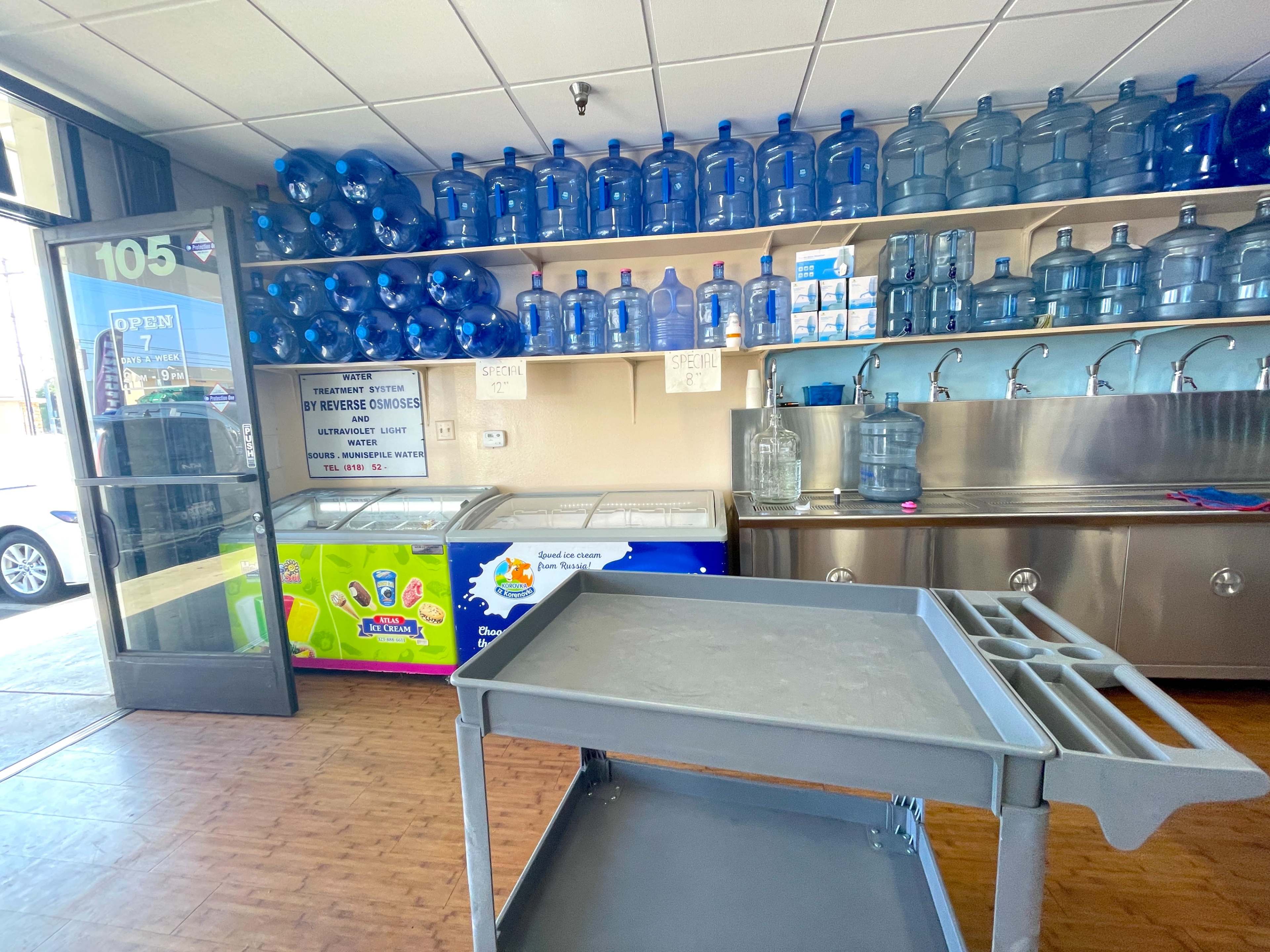A store interior features shelves lined with blue water jugs, a drink cooler, and a counter with multiple taps.