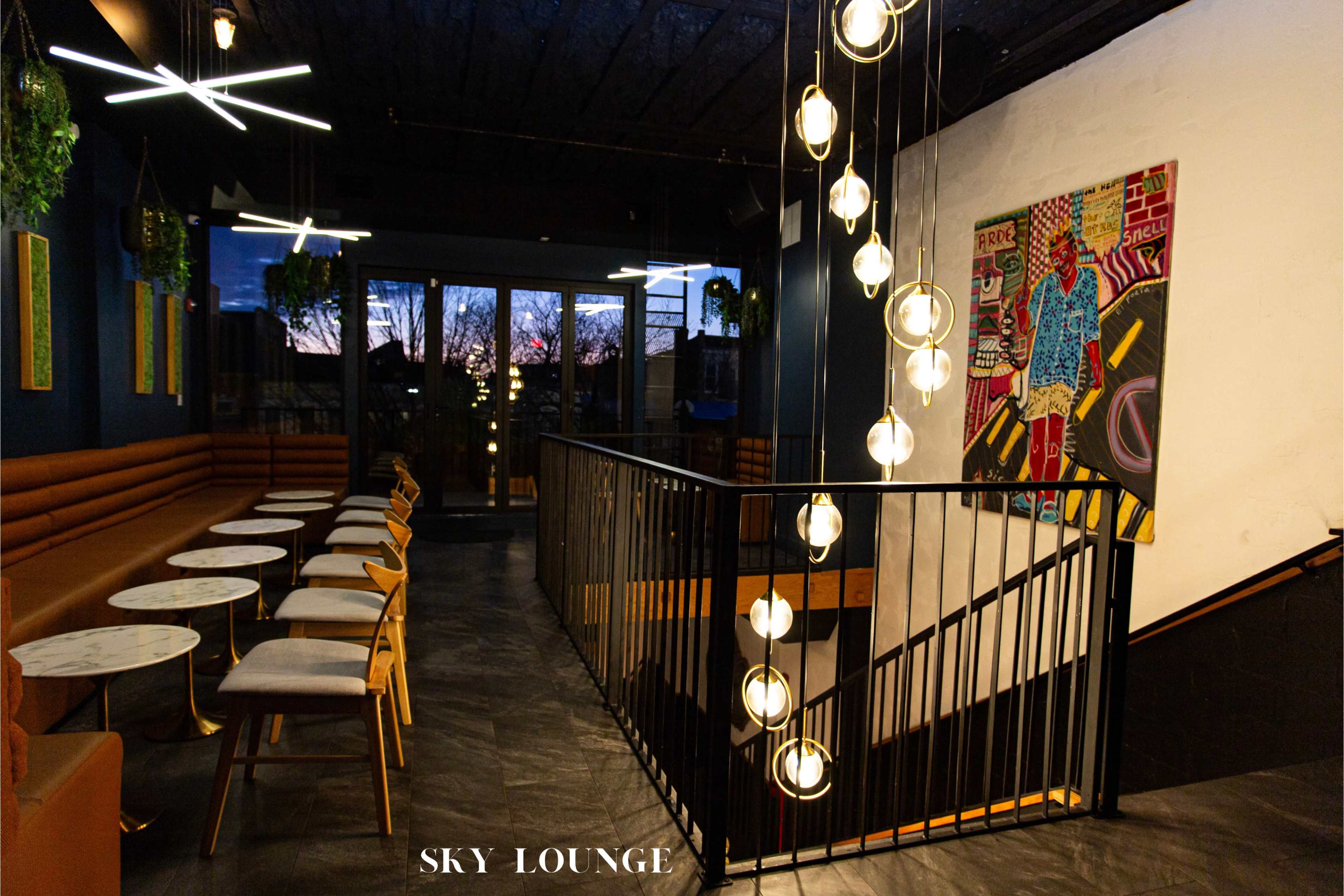 The image shows a modern lounge with seating along the walls, a decorative staircase with hanging lights, and artwork displayed on the wall.