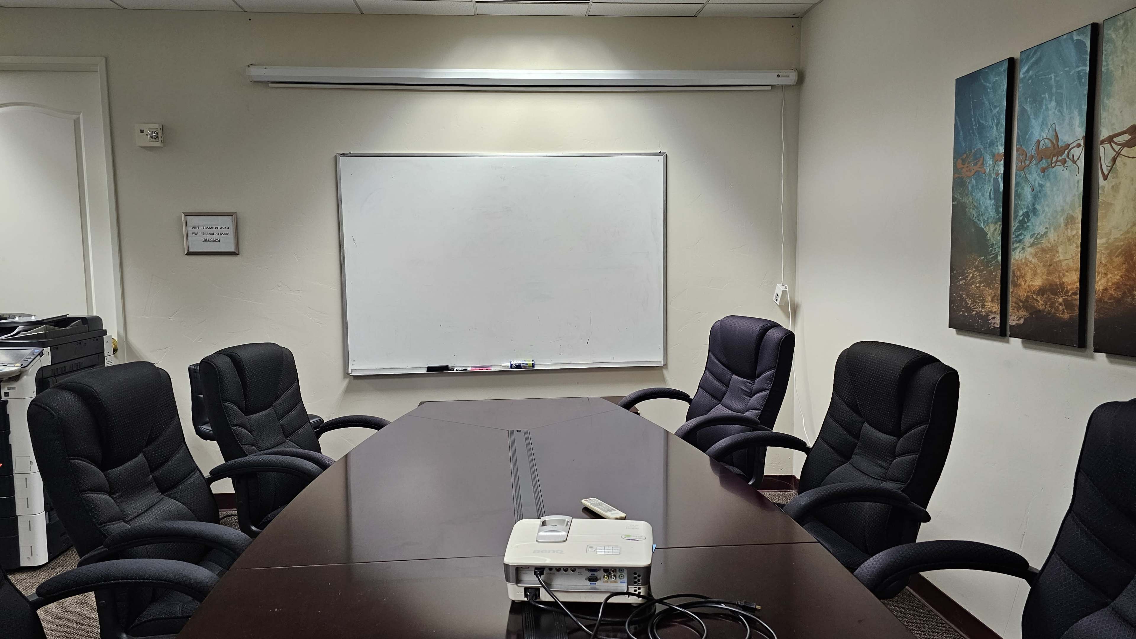 A conference room is set up with a large table surrounded by six black chairs, a whiteboard on the wall, and a projector on the table.