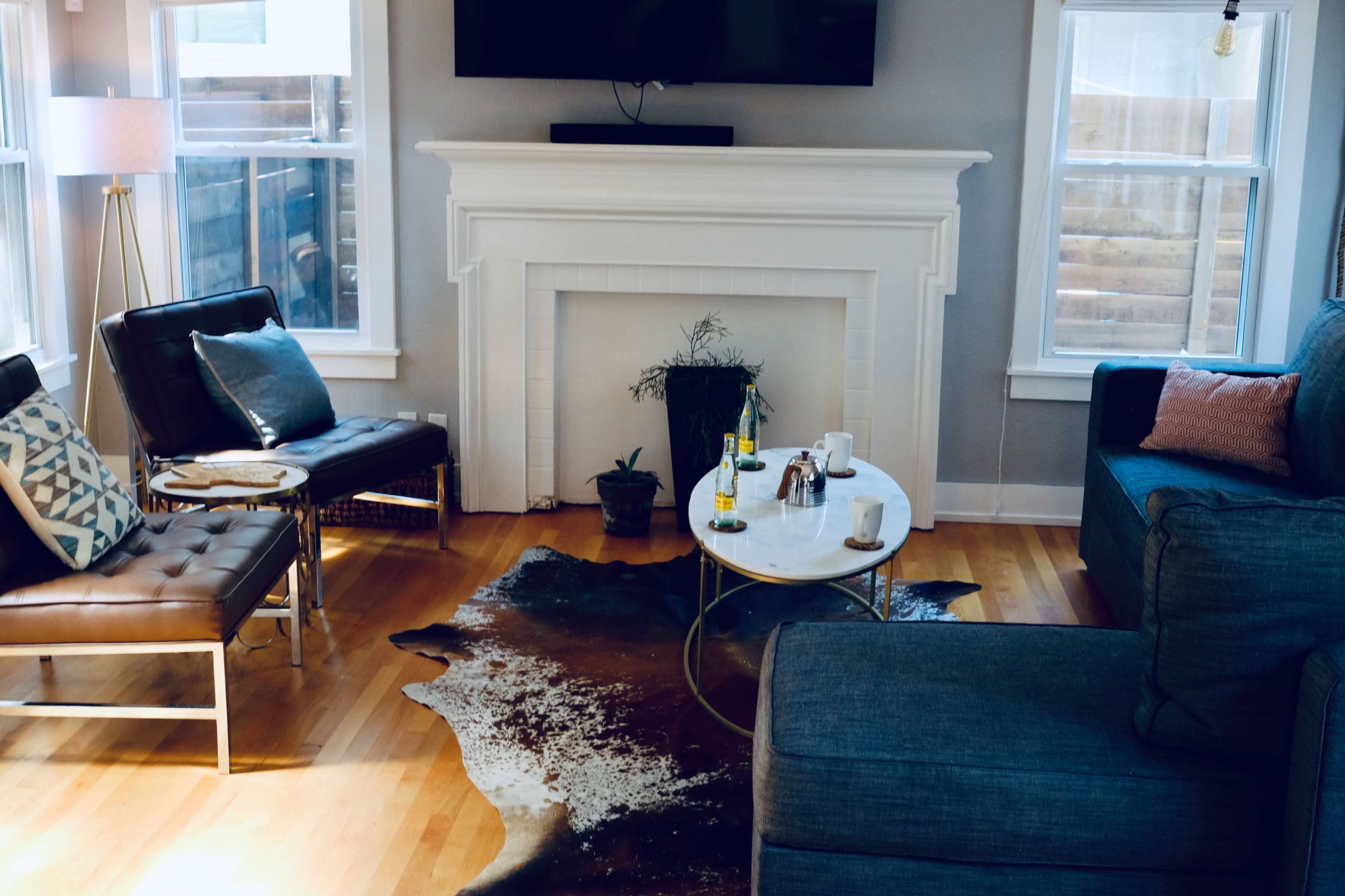 The image shows a cozy living room with a blue couch, a leather chair, a coffee table, and a decorative rug on a wooden floor, illuminated by natural light from windows.