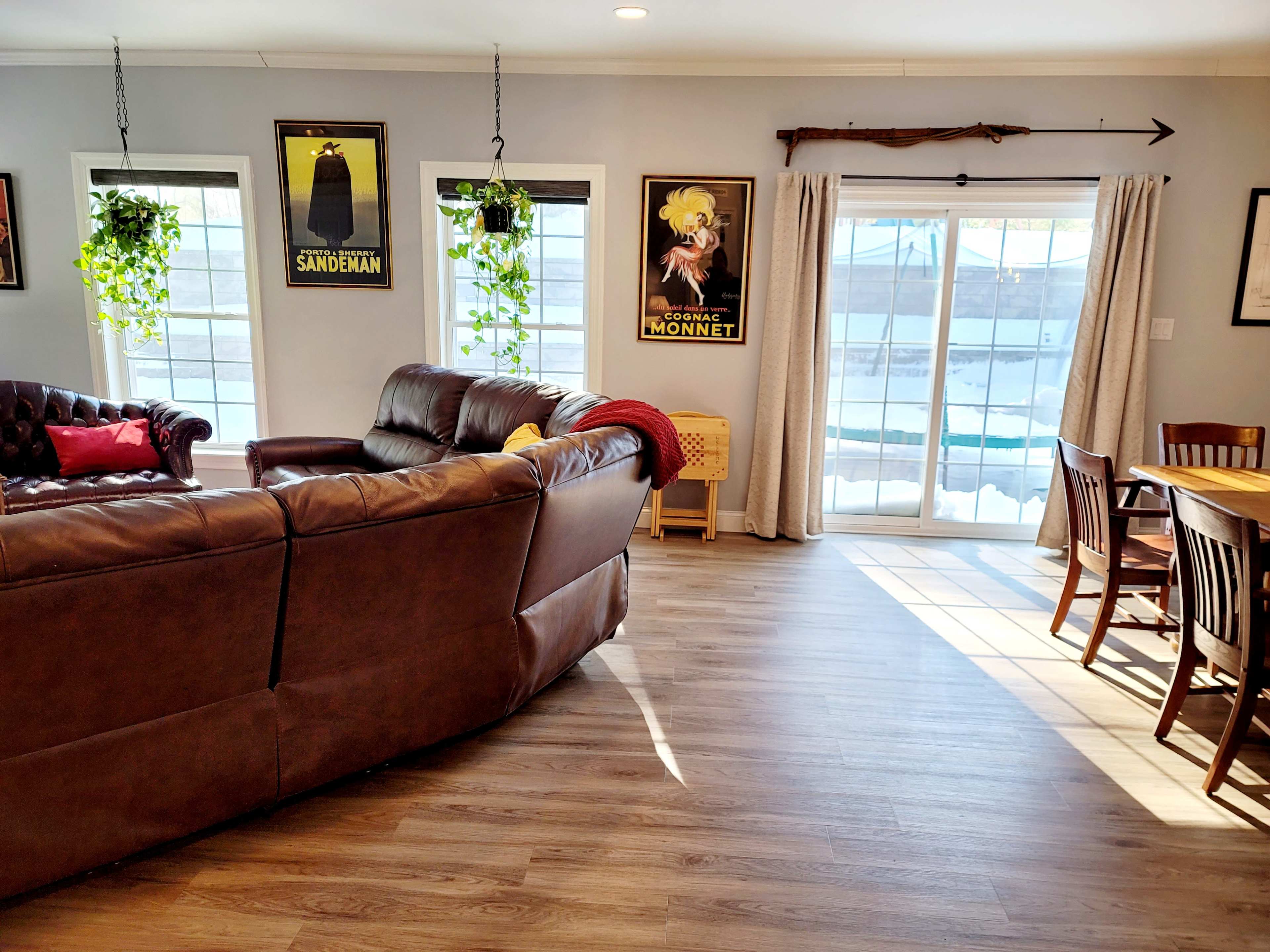 A bright living space featuring a brown leather couch, a dark wooden dining table, and large windows with potted plants on the sill.