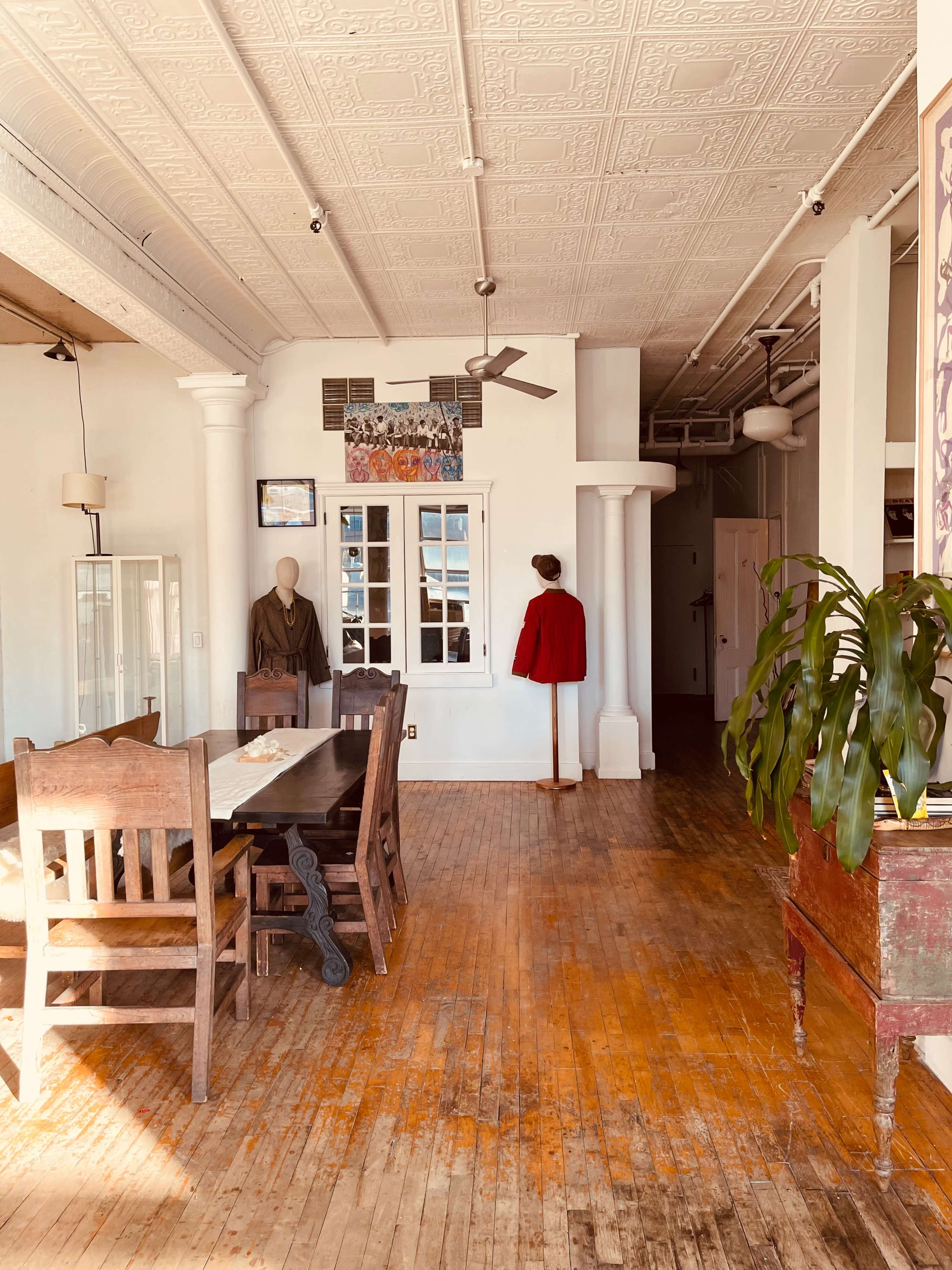 The image shows a spacious room with wooden flooring, a large dining table, a mannequin wearing a red garment, and a wall adorned with various artwork.