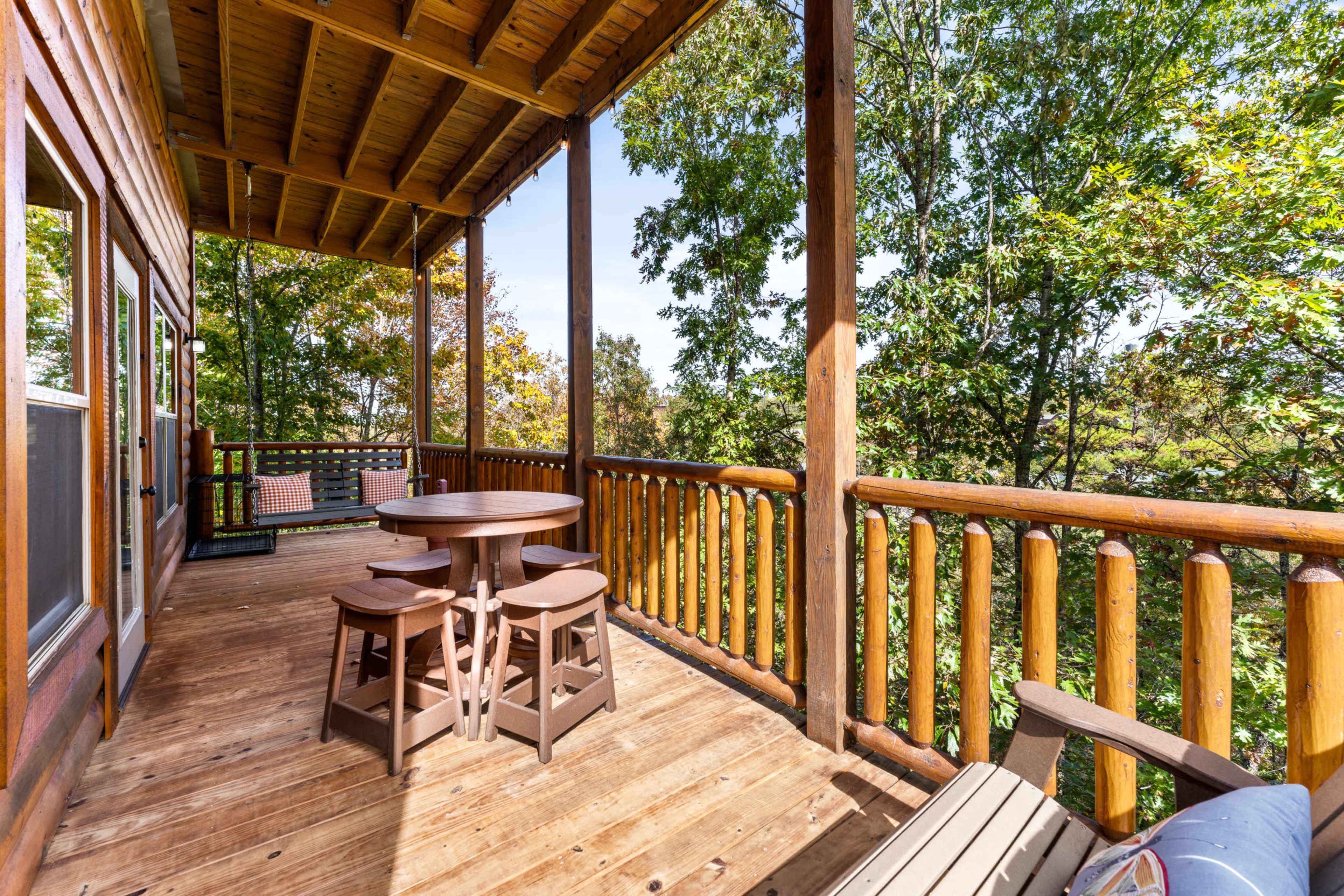 The image shows a wooden deck with a round table and stools, surrounded by trees.