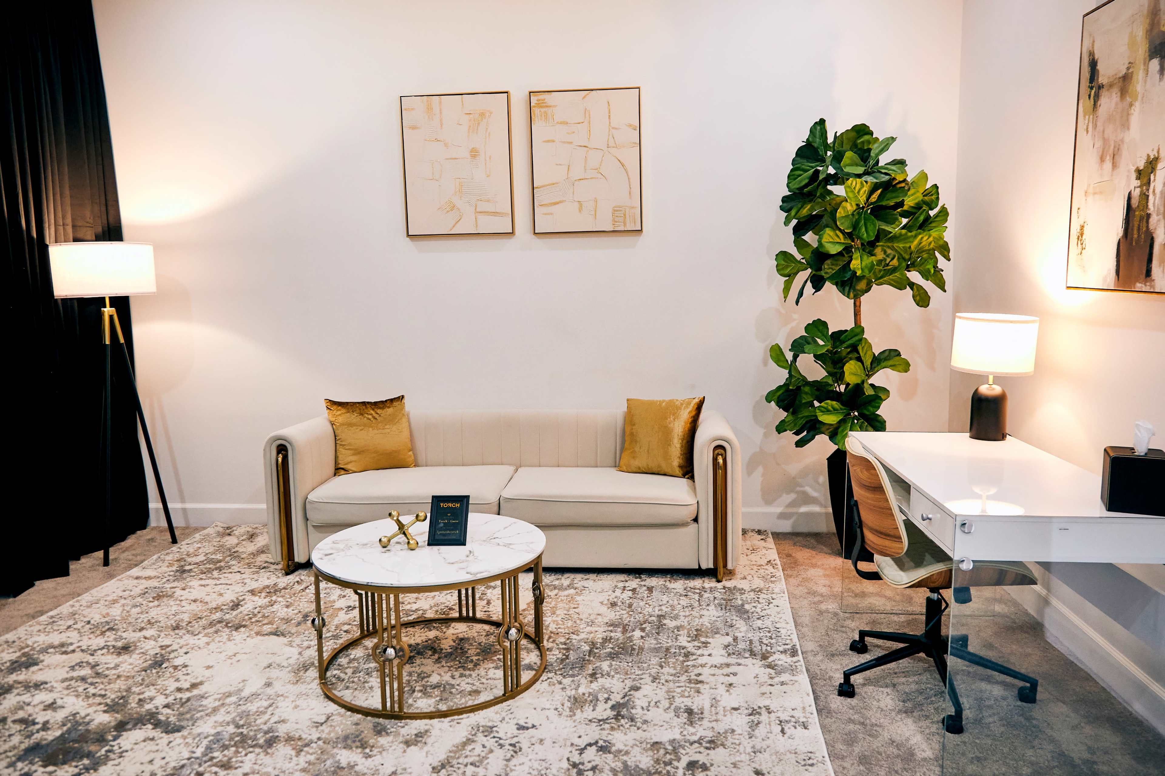 The image shows a well-decorated office space featuring a beige sofa, a marble coffee table, a desk with a chair, and two lamps, along with a large potted plant and two framed artworks on the wall.