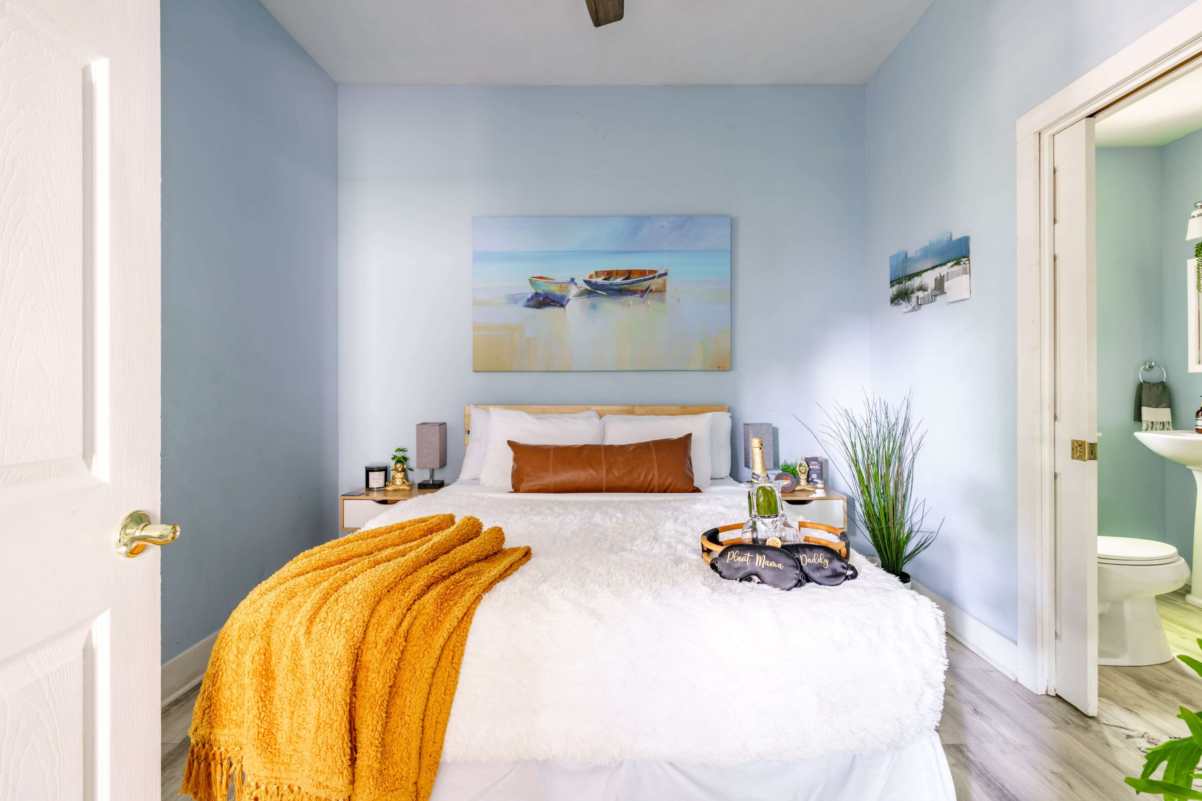 The image shows a light blue bedroom with a bed covered in a white blanket and an orange throw, complemented by a painting of boats on the wall, and a visible bathroom in the background.