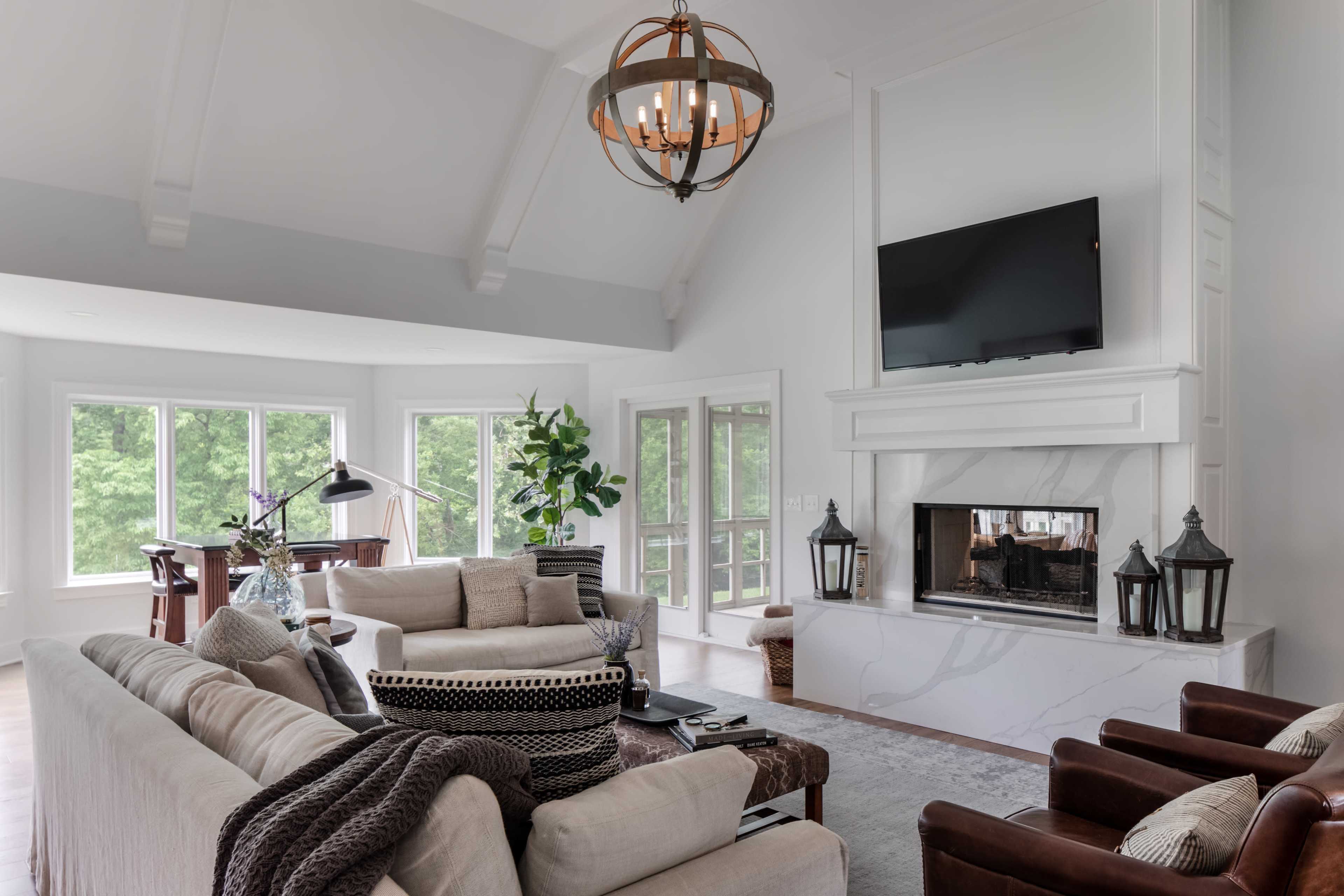 A modern living room featuring a large sofa, armchairs, a fireplace with a television above it, and large windows that provide natural light.